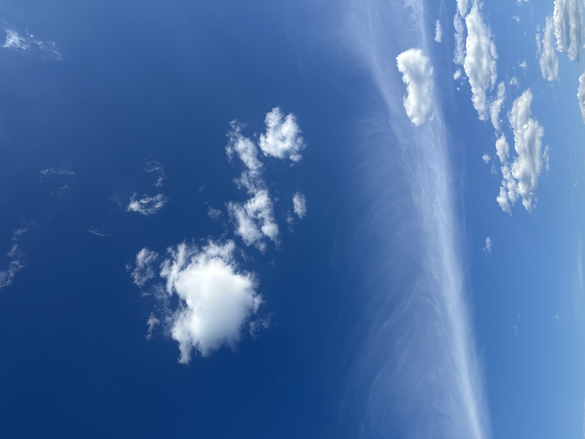 The image displays an expansive blue sky over Kharnavy, Russia. The sky transitions from a darker blue at the zenith to a lighter cerulean hue towards the lower frame. Multiple cloud types are visible. In the upper-left quadrant, a prominent, well-defined cumulus cloud exhibits a distinct, rounded form with an indentation. Scattered across the upper and mid-sections are several smaller, detached cumulus clouds. A long, attenuated band of wispy, translucent cloud material, consistent with cirrus formations, stretches horizontally across the middle portion of the sky, extending from left to right. A cluster of additional cumulus clouds is present in the lower-right section. No ground features, structures, or individuals are visible within the frame.