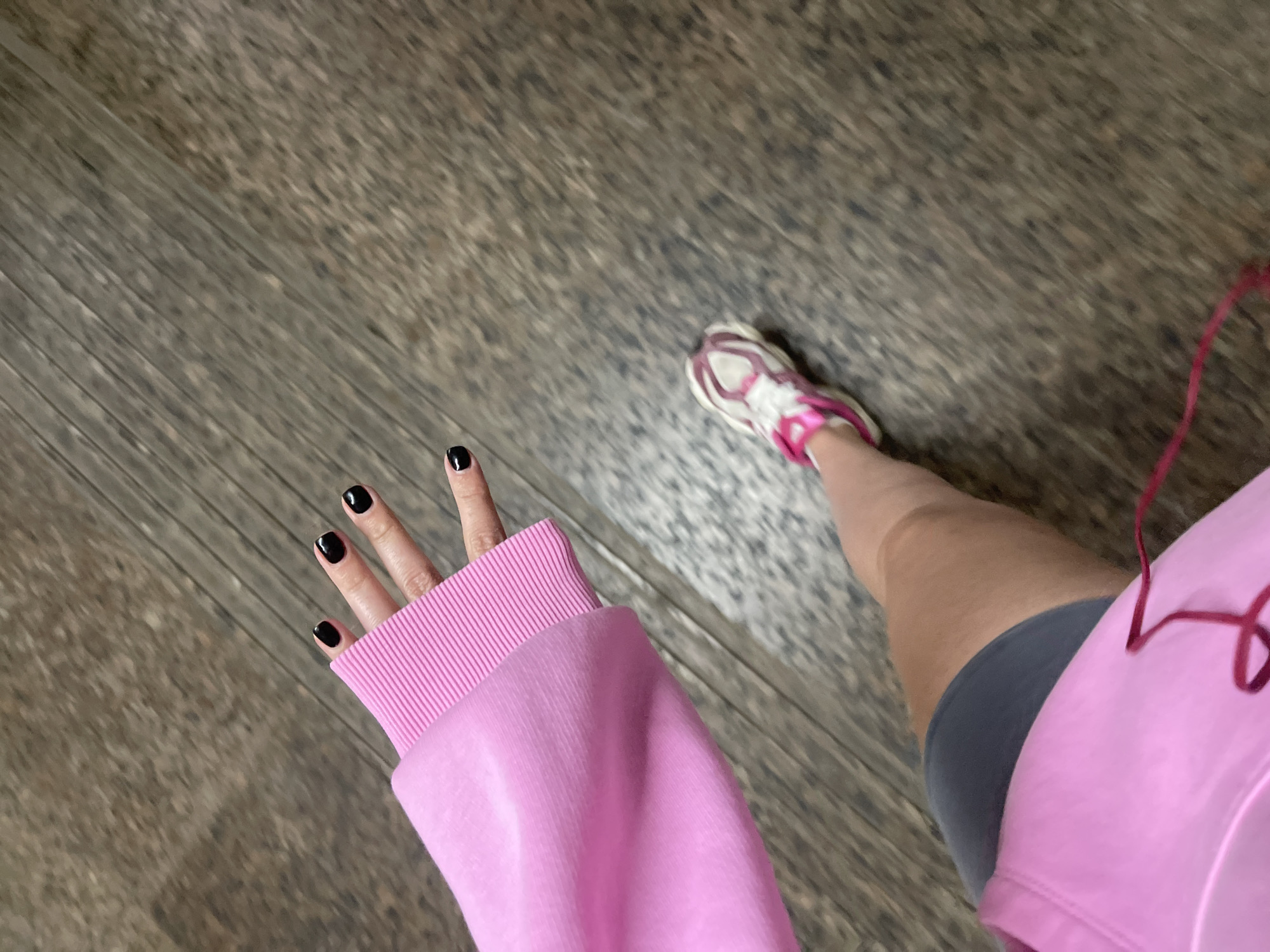 A first-person, eye-level shot shows a person on a weathered, wood-look floor. The subject's left hand, adorned with black nail polish, extends into the frame, partially covered by a pink sweatshirt cuff. The person's leg, clad in gray shorts, is visible, leading to a pink and white running shoe. A red wire suggests headphones or earbuds. The setting appears to be indoors, possibly a home or gym. There is no evidence to suggest this image was taken in Moscow, Russia.