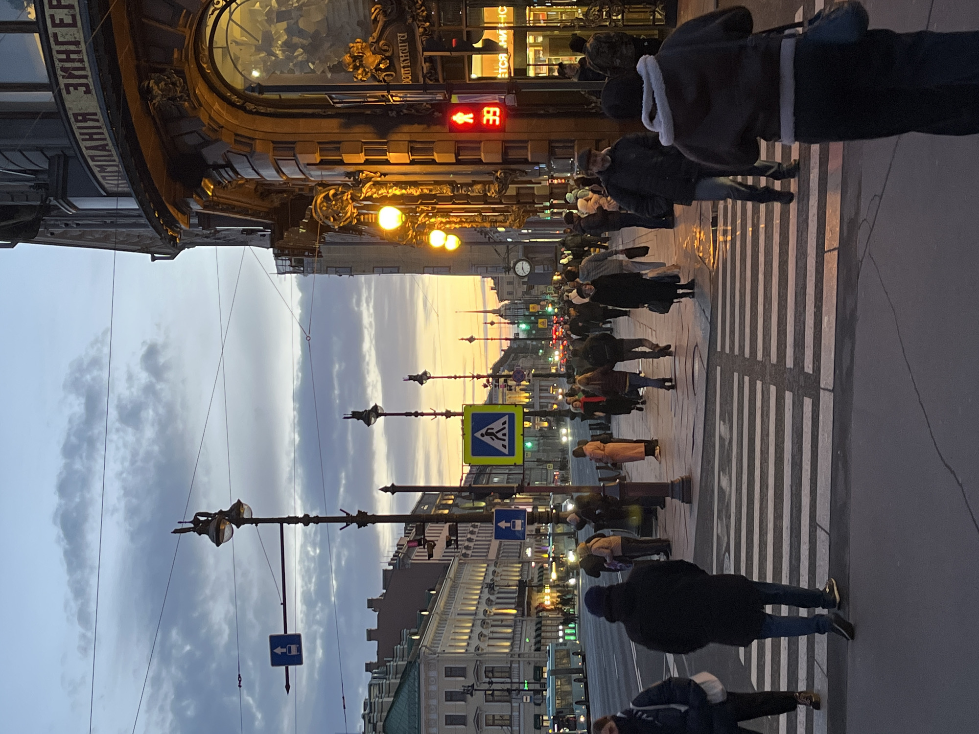 The image shows a street scene in Saint Petersburg, Russia, at dusk. People are crossing a zebra-striped pedestrian crosswalk. On the left, a large ornate building with Cyrillic text is visible. Street lamps line the street leading to the distant horizon. The sky is cloudy with a hint of sunset. Traffic lights are displayed above the street. Pedestrian and directional signs are also present along the street.