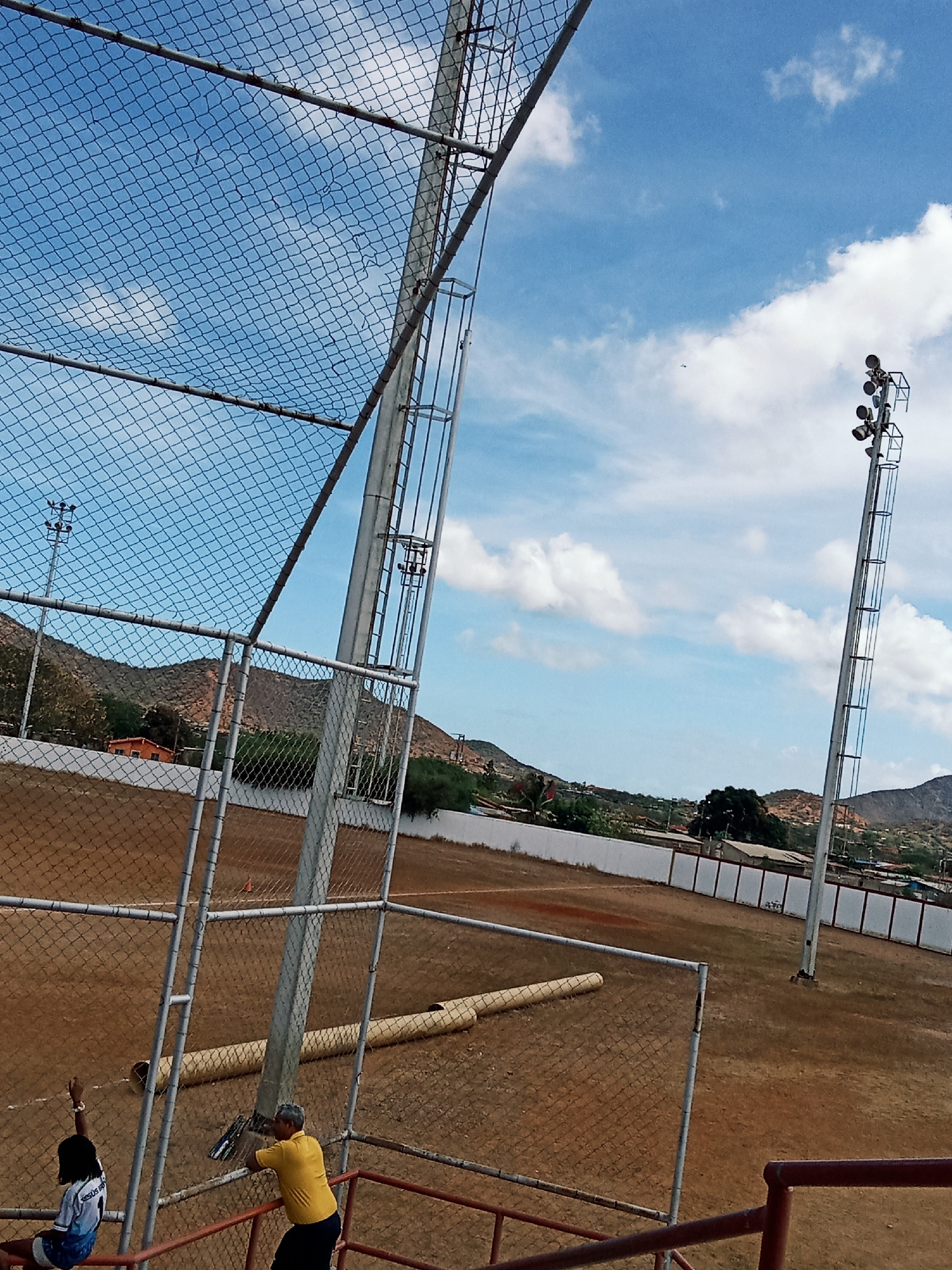 This outdoor image depicts a baseball field located in Juan Griego, Venezuela. A chain-link fence encloses the field. Two light towers stand on the field. A partial view of two individuals can be observed in the foreground: one sitting, one leaning against a fence. A long, cylindrical object lies on the field near the fence line. Mountains are visible in the background, under a blue sky with white clouds. The ground is dirt and dry grass. A white wall with intermittent dark vertical markings surrounds the outer edge of the field.