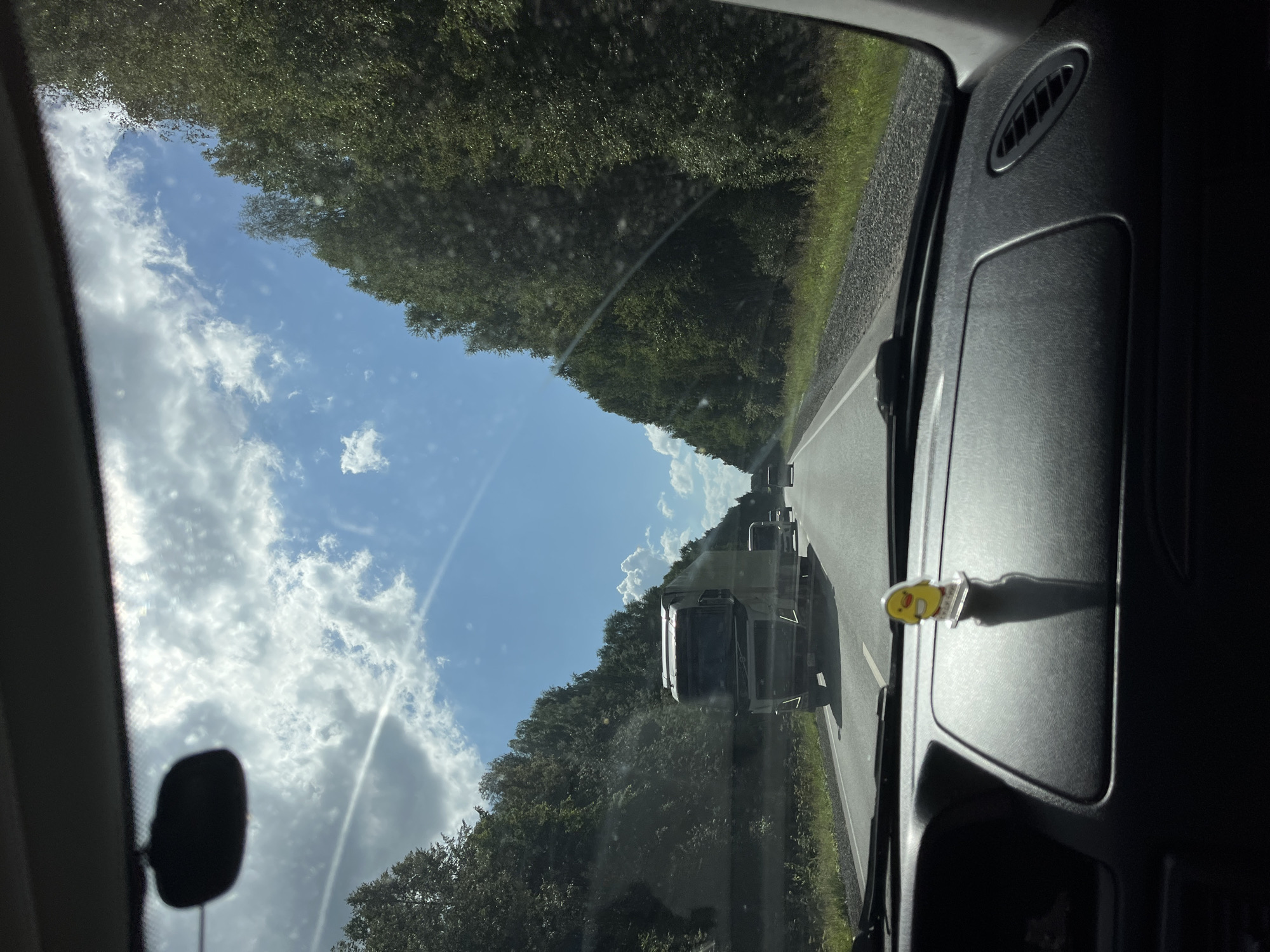 The image shows a view from inside a car driving on a highway in Uspenskii, Russia. A large semi-truck is visible in the distance on the right lane of the highway, followed by other vehicles. The highway is lined with dense green trees on both sides, and the sky is blue with scattered white clouds. The dashboard of the car is in the foreground, with a small yellow figurine of a chick placed on it. The overall scene depicts a sunny day on a road trip.