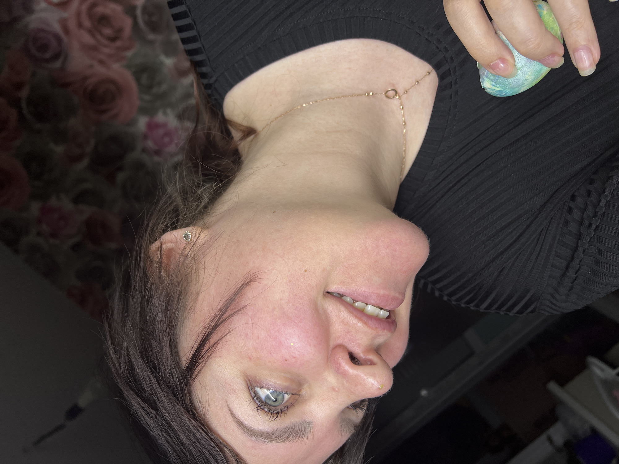 A woman with fair skin and light eyes is photographed indoors from a close-up, slightly upward angle. She is wearing a black ribbed top and a delicate gold necklace. Her hair is dark brown with some light brown highlights. She has a stud earring in her left ear. She is holding a blue and green makeup sponge in her right hand. The background behind her consists of a floral print wallpaper and a dimly lit area with what appears to be furniture or equipment. There is no evidence to suggest this image was taken in the city of Noviy, Russia.