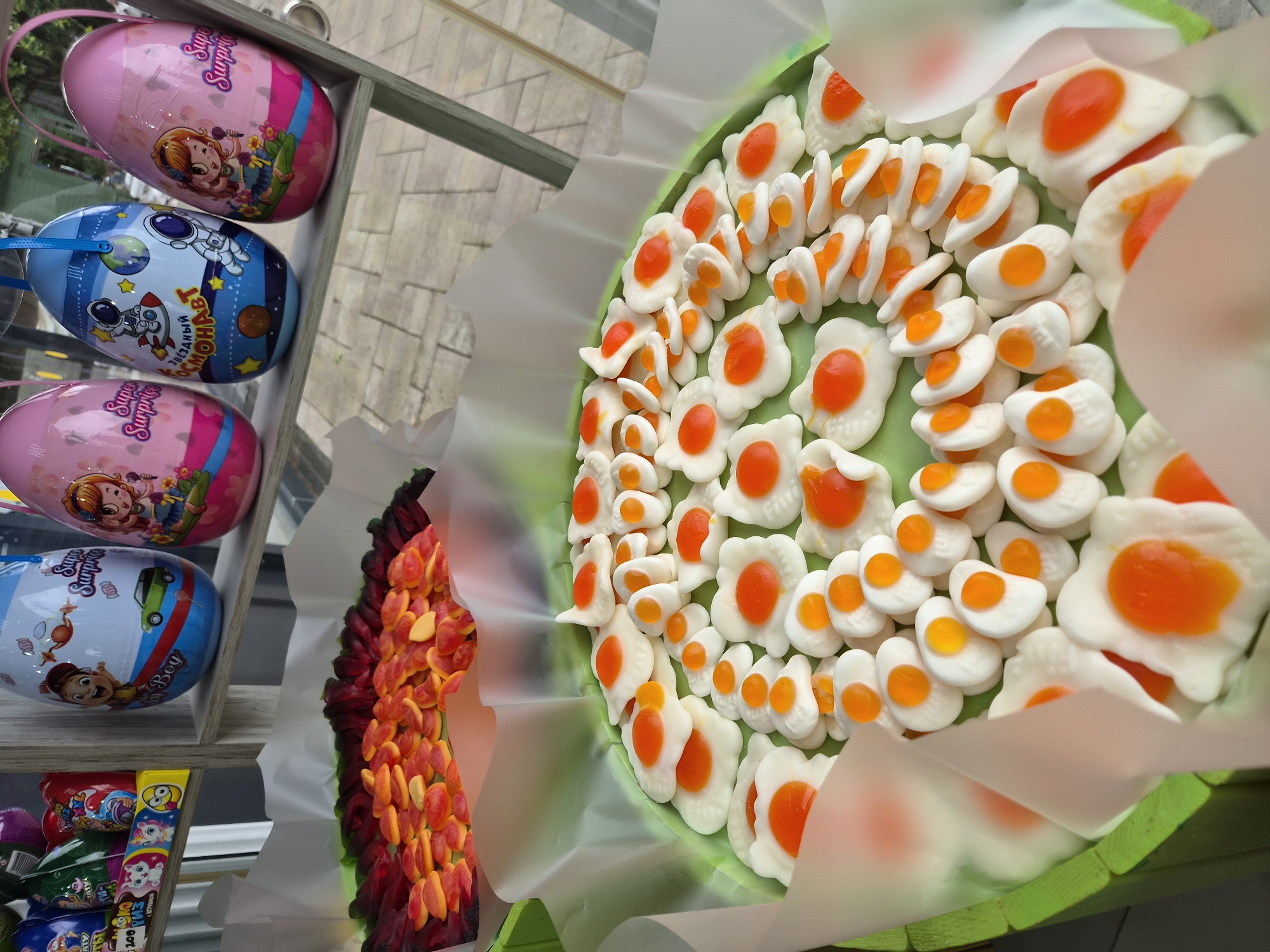 A close-up captures a display of confectionery and treats in what appears to be a shop or vendor stall, potentially located in Sochi, Russia, based on the Cyrillic text present on one of the items. The focal point is a circular arrangement of gummy candies shaped like fried eggs with white "egg whites" and orange "yolks," presented in a decorative green container. To the left of the candy eggs is a collection of gummy candies with a gradient from red to orange.
On a multi-tiered wooden shelf to the far left, there are various items, including plastic egg-shaped containers with children's cartoon characters and labels like "Super Surprise" written in English, as well as Cyrillic script, which is suggestive of a Russian origin. Other packages of candies and sweets are also visible on the shelf. The arrangement suggests a display intended to attract children or those with a fondness for confectionery.
