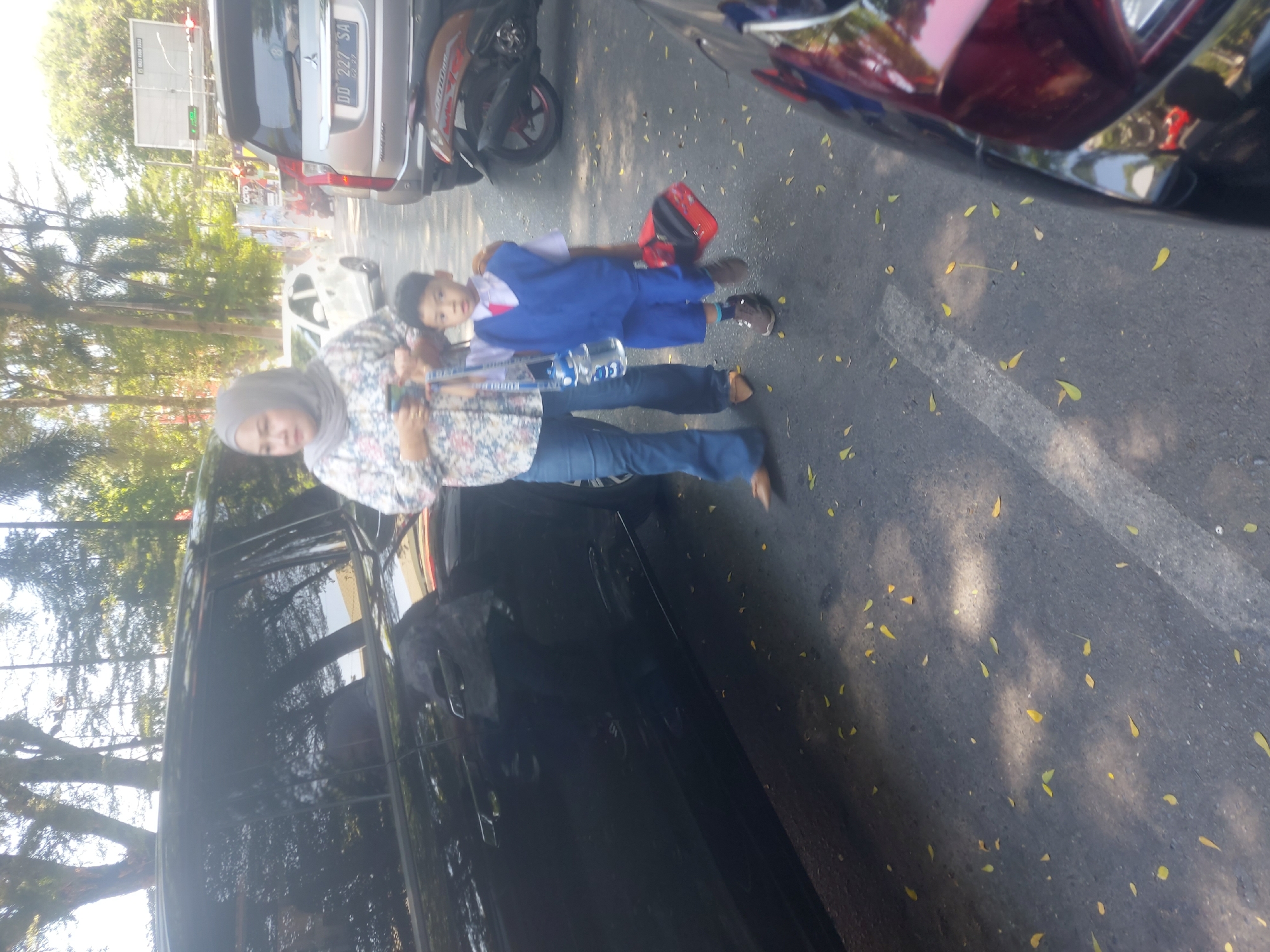 This is a medium shot, eye-level, vertical, outdoor photograph taken during the day. In the foreground, a black SUV is partially visible on the left side of the frame. In the middle ground, a woman wearing a hijab and a floral-patterned shirt, and blue jeans is walking. She is holding a light blue water bottle with a handle. Beside her, a young boy in a school uniform consisting of a white shirt, blue vest, blue shorts, and red tie is walking, carrying a red bag. He is also wearing brown shoes. Behind them, a silver SUV with license plate "DD 227 SA" is parked. A brown motorcycle is parked behind the silver SUV. The background consists of trees and a blue sky. The scene appears to be in a city environment, possibly in Makassar, Indonesia, given the license plate format. The ground is paved and scattered with yellow leaves. The overall lighting suggests a sunny day.