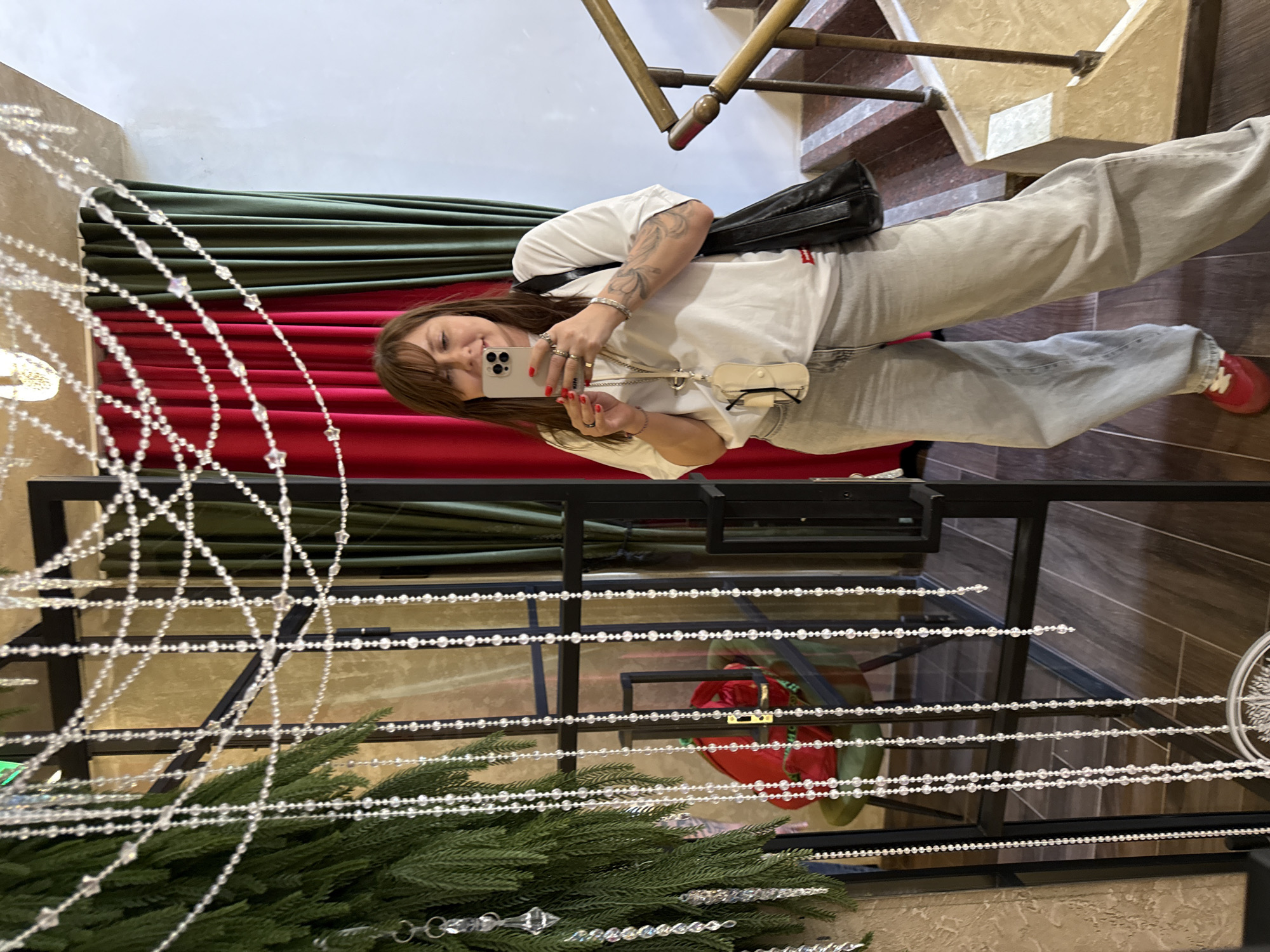 A woman with brown hair is taking a selfie in what appears to be a hotel lobby or a similar establishment in Sochi, Russia. She is wearing a white t-shirt, light gray wide-leg jeans, and red shoes. She has a black bag slung over her shoulder and glasses hanging from a lanyard around her neck. The background features a staircase with a gold-colored railing, plush red and green velvet curtains, and a decorative element of hanging clear beads and greenery. A chair with a red object is visible in the reflection of a mirrored surface.