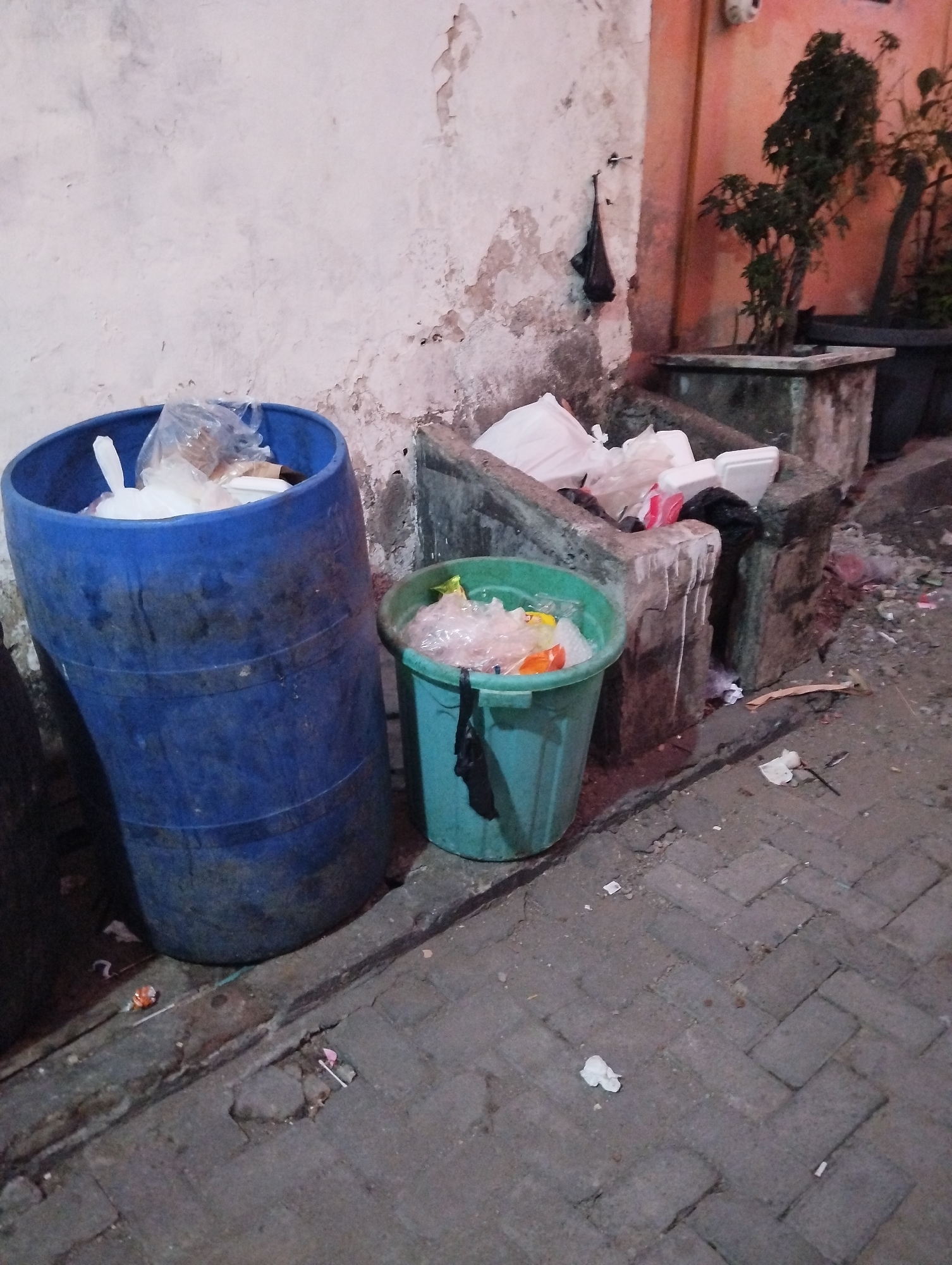 The image depicts an outdoor scene in Kepuh Kiriman, Indonesia, likely a public alley or street corner. Two large blue and green plastic trash bins are prominently featured, both overflowing with garbage, primarily plastic bags and white packaging materials. To the right of the bins, there's a concrete structure that appears to be a planter or another receptacle, also containing refuse and some potted plants. The ground is paved with interlocking bricks, and litter is scattered around the bins and the concrete structure. A distressed wall, possibly of a building, forms the backdrop, with peeling paint and rough texture. The lighting suggests it might be dusk or nighttime.