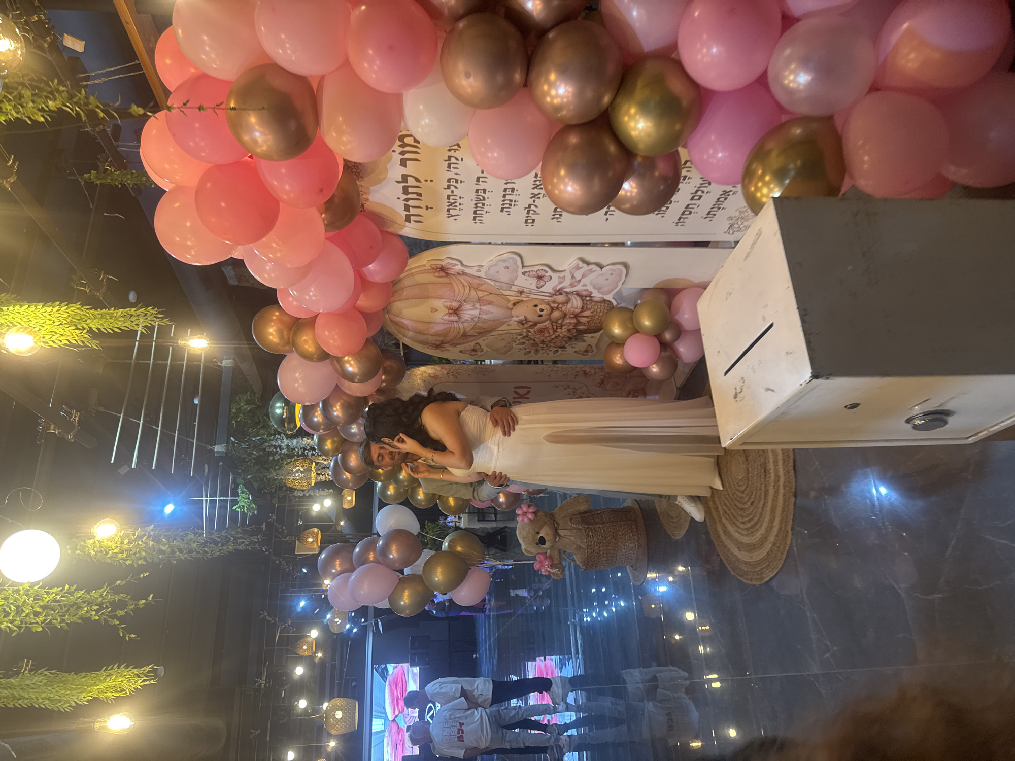 The image depicts a celebratory indoor event, likely a baby shower or birthday, with a prominent display of pink and gold balloons. In the foreground, a couple is embracing, with the woman wearing a long, strapless white dress and the man in a light-colored suit. Beside them, a teddy bear sits in a woven basket adorned with small balloons. Behind the couple, a large decorative cutout features a hot air balloon and a teddy bear, alongside a sign with Hebrew text. The venue features dark floors with reflective surfaces, hanging greenery, and industrial-style lighting. In the background, several individuals are visible, and a large screen displays abstract imagery. The overall atmosphere is festive and intimate. The sign with Hebrew text indicates the event is likely in Migdal HaEmek, Israel, a common practice for such celebrations.