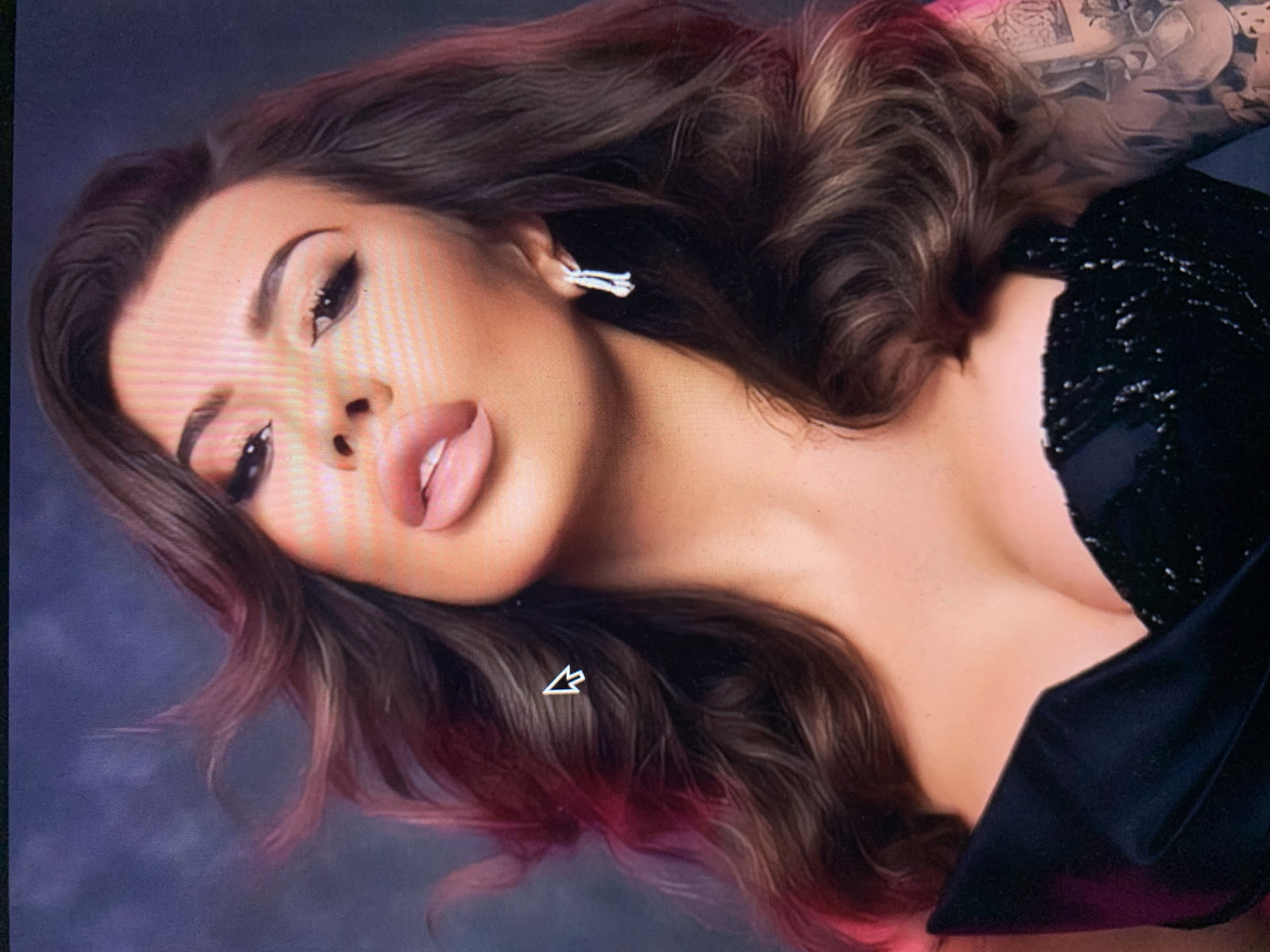 An over-the-shoulder, medium shot of a woman with dark, wavy hair and full lips. She is wearing a black, strapless dress with a sequined bodice. She has large, dark eyes and her hair is blowing to the left. She is wearing a small, silver earring in her right ear. The background is a blurred, dark blue-gray color with a splash of pink light on the left side. There is a computer cursor in the center left of the image.