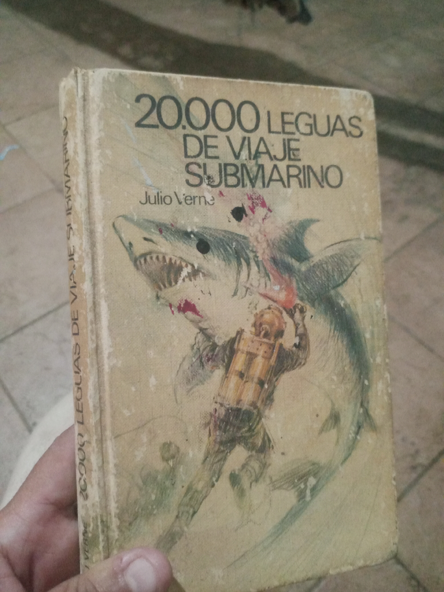The image shows a vintage book with a worn cover. The title on the cover reads "20,000 LEGUAS DE VIAJE SUBMARINO" which translates to "20,000 Leagues Under the Sea." The author's name, "Julio Verne," is also visible. The illustration on the cover depicts a shark attacking a diver in a vintage diving suit. The book is held by a person's hand. The background appears to be an indoor setting with a tiled floor. There is no information in the image to determine its location in Santa Ana, Venezuela.