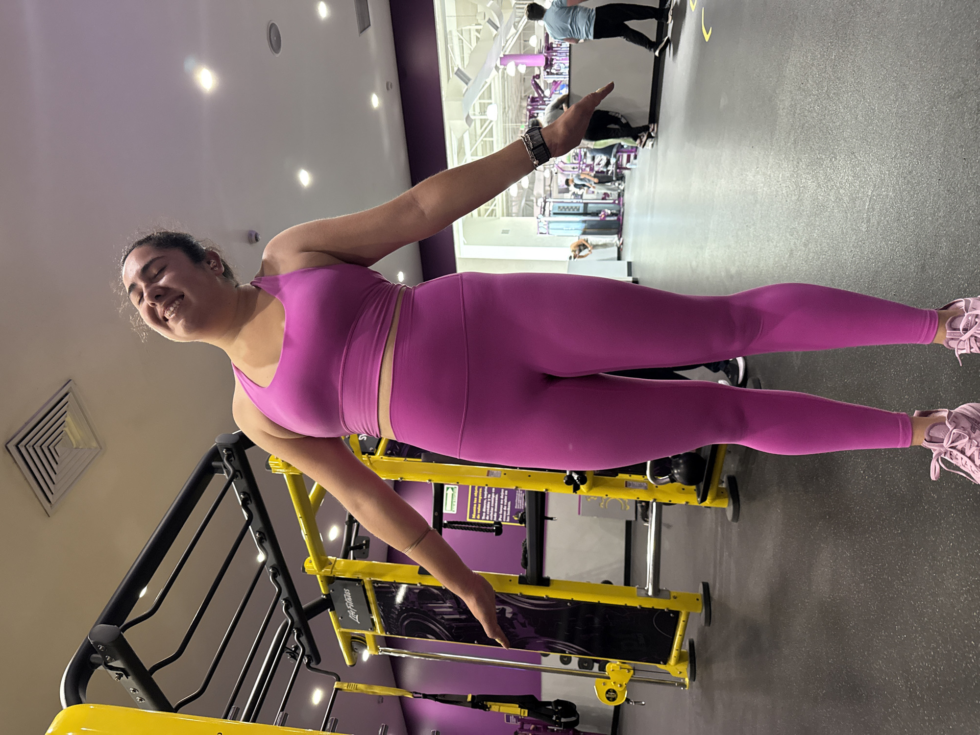 A woman in a pink workout outfit is standing in a gym with her arms outstretched, facing the camera. She is smiling. The gym has yellow and black Life Fitness equipment. Other people can be seen in the background. The gym's interior is lit with overhead fluorescent lights. It is not possible to confirm that the location is Puebla, Mexico, based on the provided image.