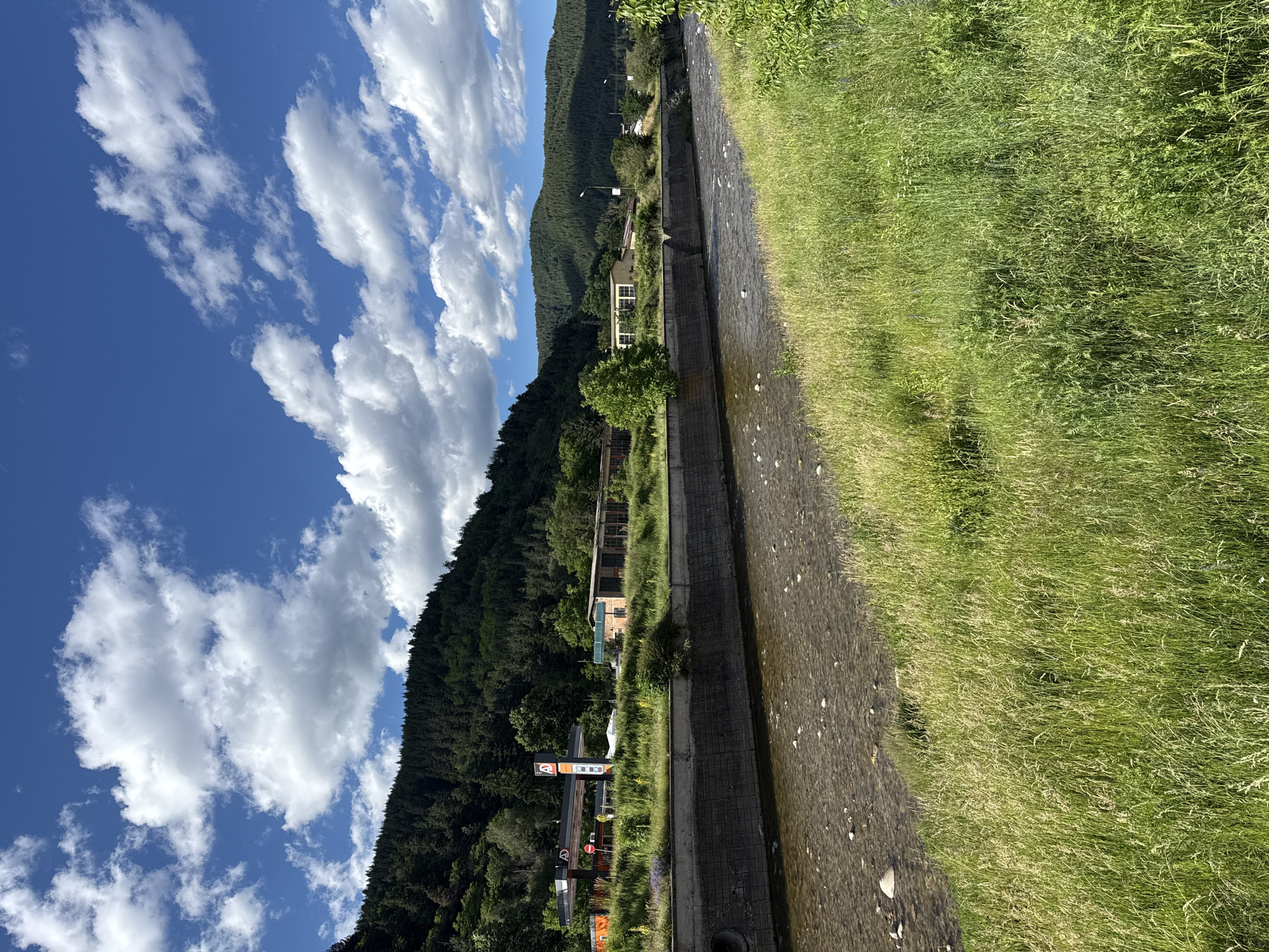 The image shows a natural scene with a river flowing through a landscape in Yakoruda, Bulgaria. The river is shallow and appears to have a rocky bottom. On the left bank of the river, there is a concrete embankment with a gas station and some industrial buildings behind it. The background is dominated by a densely forested hill, with more hills visible in the distance. The sky is blue with scattered white clouds. The foreground features a grassy embankment with tall grass. The overall impression is one of a rural or semi-rural environment, likely on the outskirts of the town.