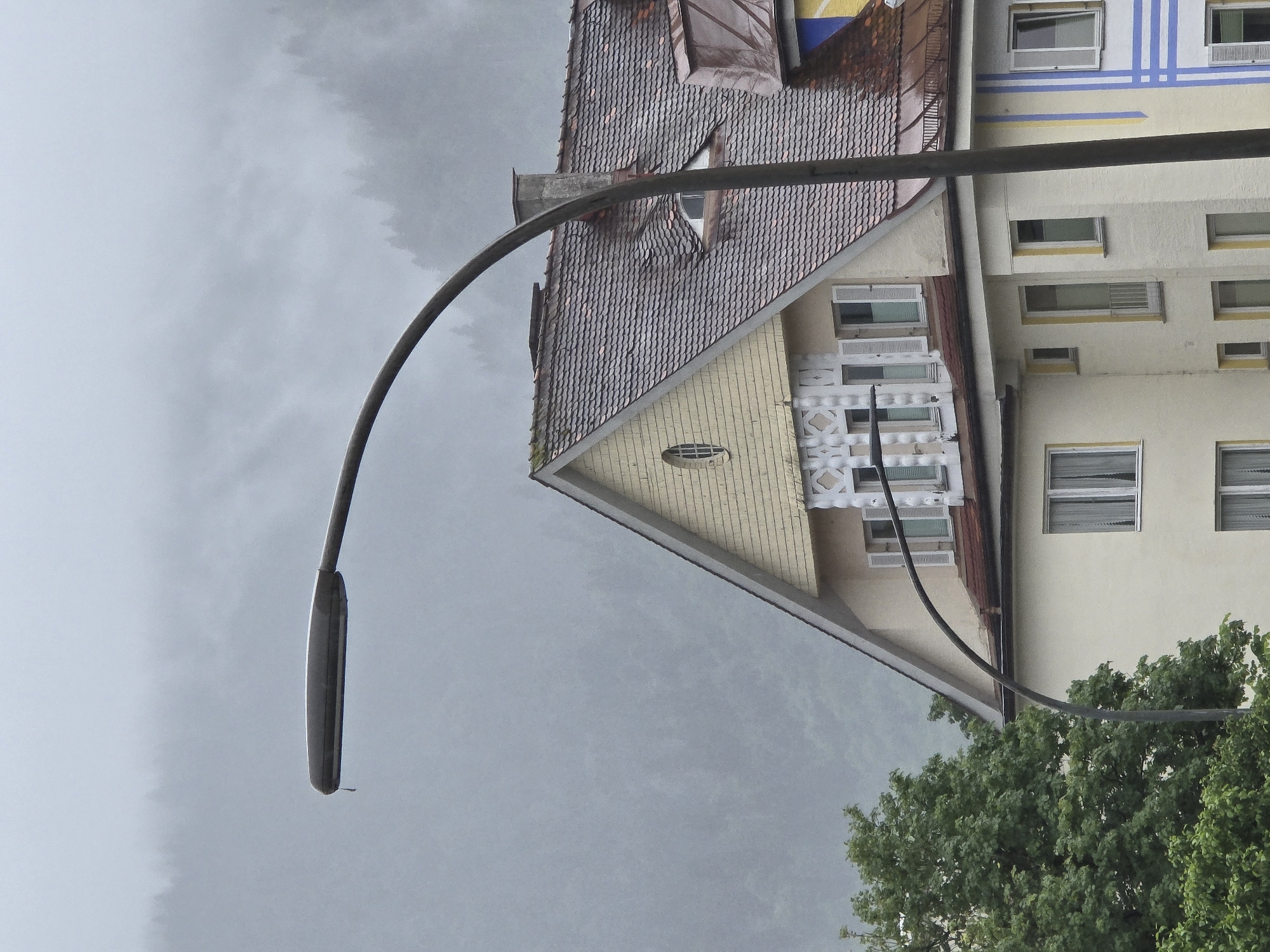 This media file captures an outdoor scene with a focus on a building and a streetlamp against a cloudy sky. The image is oriented vertically, with the top of the frame showing a muted, overcast sky. A prominent curved black streetlamp extends from the bottom left of the frame upwards, crossing the middle.
The main subject is a building with distinct architectural features. The left side of the building has a steeply pitched, dark-tiled roof, appearing older, with a dormer window. Adjacent to this, a section of the building is painted a light yellow, with horizontal siding and decorative white latticework around some of the windows. Further to the right, the building's facade is painted a pale cream color. This section features several windows of varying sizes, some with white shutters. Blue and yellow horizontal stripes are visible across the top of this cream-colored section, suggesting it might be a more modern addition or decoration.
At the bottom right of the image, lush green foliage from trees is visible, partially obscuring the base of the building and suggesting an urban or suburban setting with greenery.
The weather appears to be overcast and possibly damp, indicated by the muted lighting and the texture of the sky. The scene does not depict any people or specific activities. The location context suggests Garmisch-Partenkirchen, Germany, though no explicit text or signs are visible to confirm this. The overall mood is somewhat subdued due to the cloudy weather and the lack of activity.