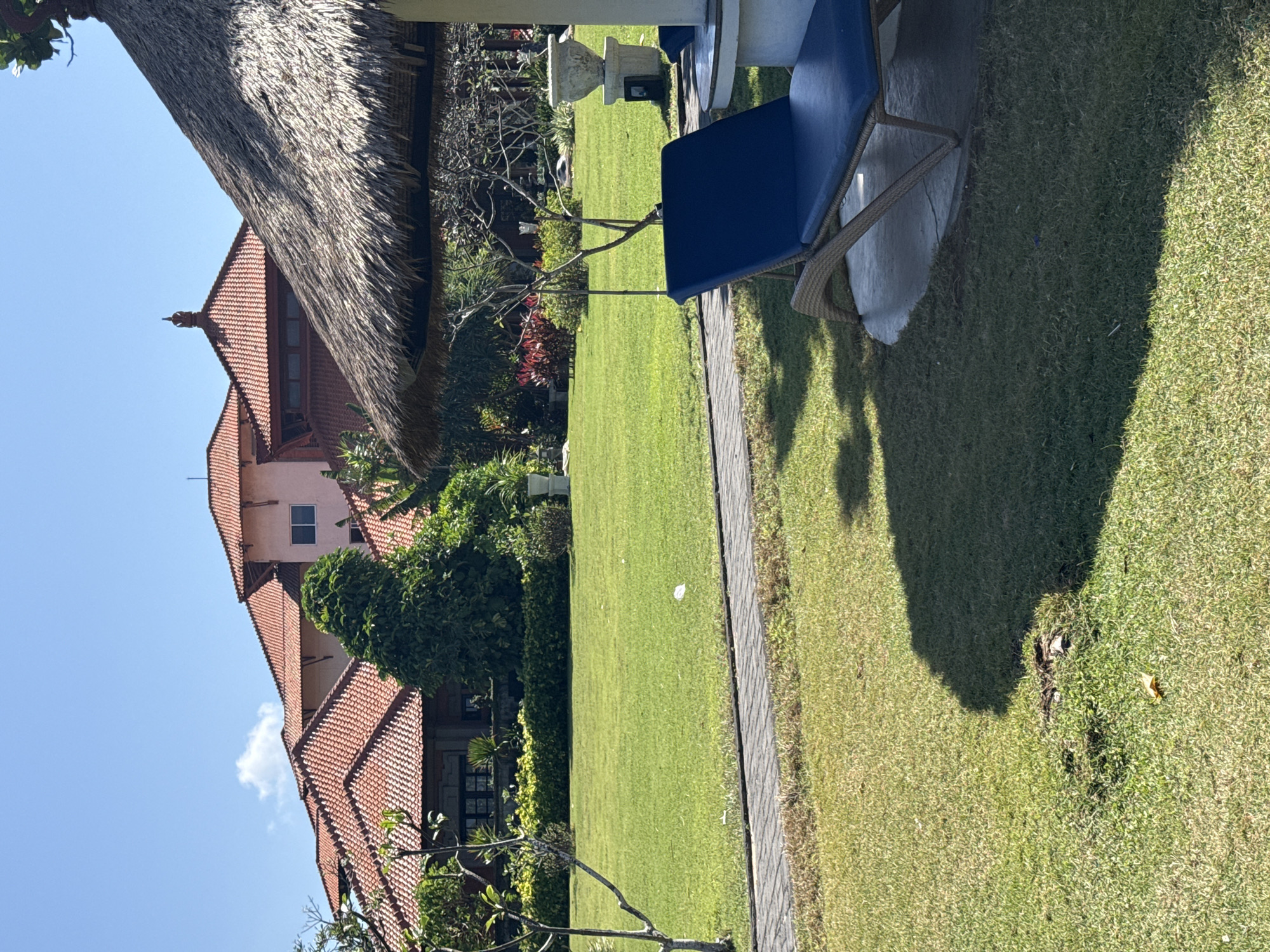 The image depicts a resort lawn in Kuta Selatan, Indonesia. A blue chaise lounge sits on a paved area on the right side of the frame, partially shaded by a thatched-roof umbrella. In the background, a multi-story building with terracotta tiled roofs is visible. The lawn is well-maintained and bordered by a paved walkway. Trees and shrubs are scattered throughout the grounds, and the sky is clear and blue. The overall scene suggests a tranquil resort setting.