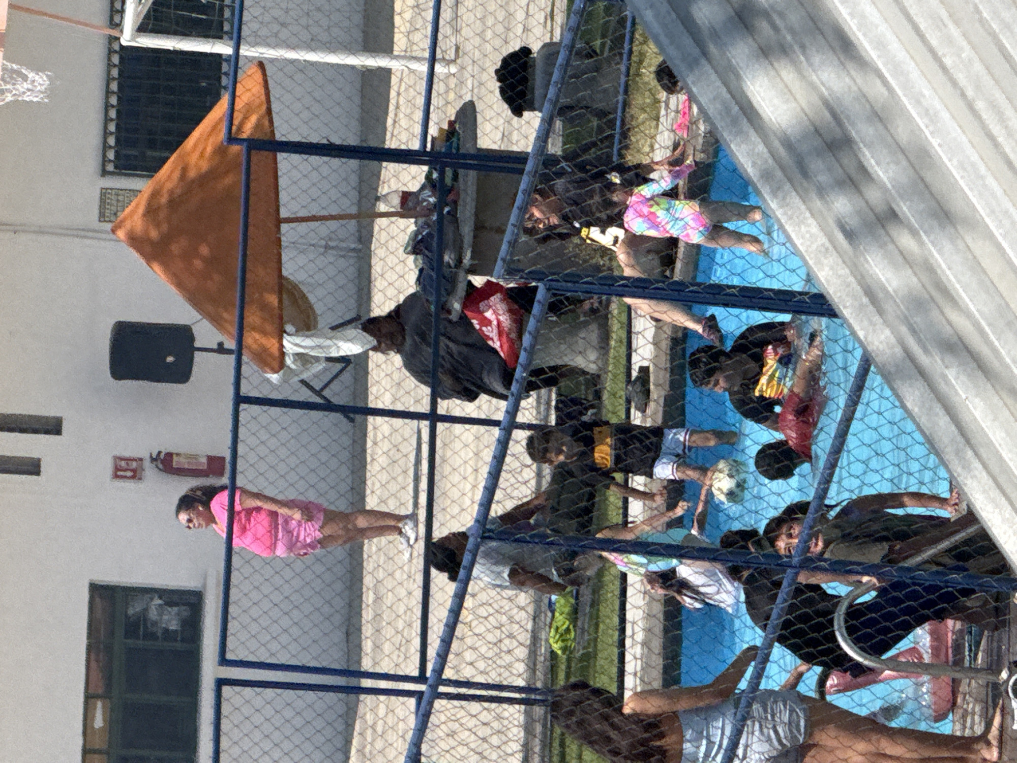 Observed from an elevated position through a chain-link fence, the image captures a group of individuals around a pool, located in what appears to be a courtyard setting. Several people are in the pool, engaging in various activities. Others are positioned around the pool's edge. A woman in pink stands on the building's periphery. In the background, a white building with a window, a fire extinguisher, and an orange triangular sunshade are visible. The location is identified as Puebla, Mexico, based on context.