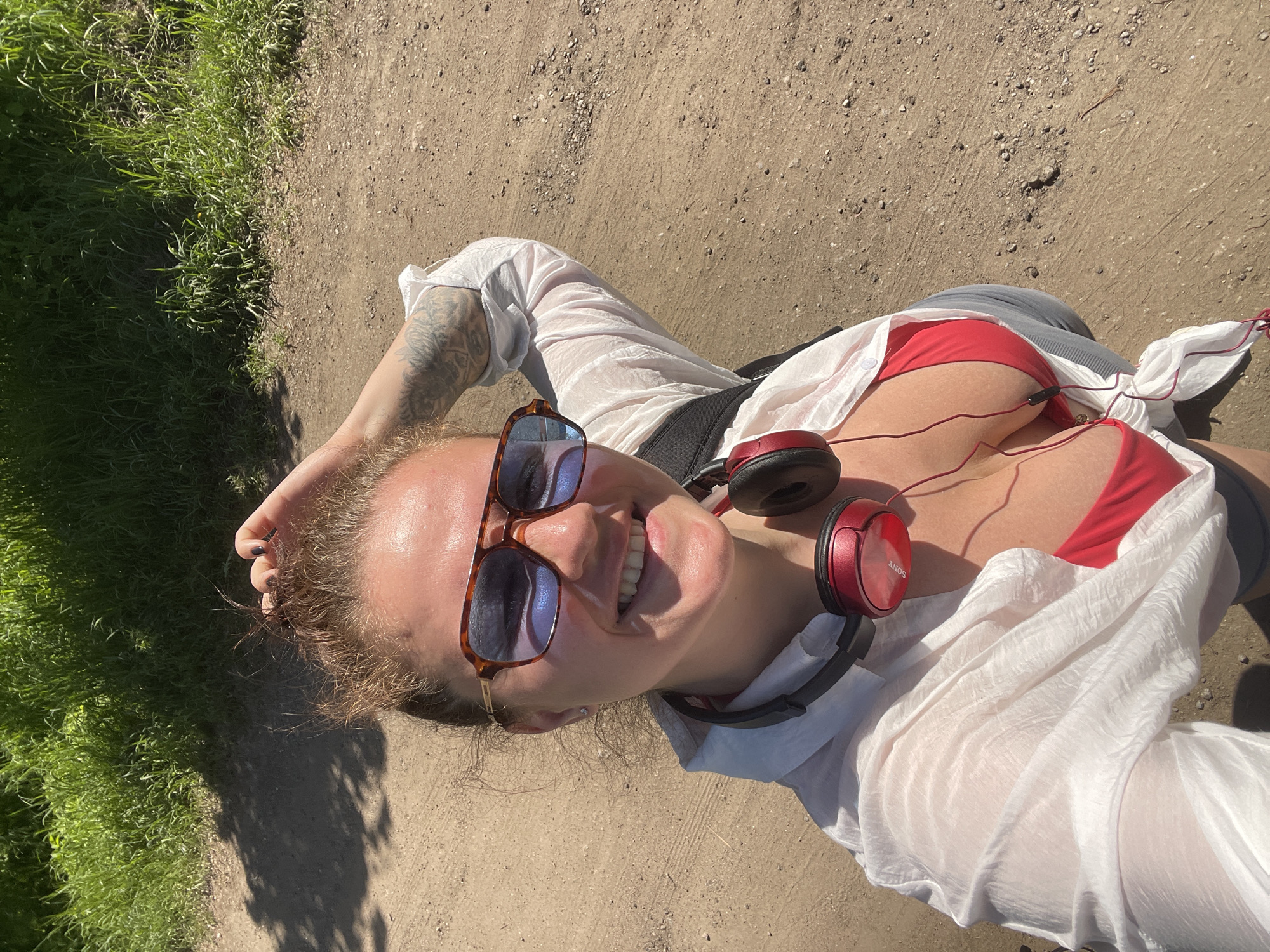 A woman is lying on a dirt path near grass. She is wearing sunglasses, a white button-down shirt over a red swimsuit top, and red headphones around her neck. She has a tattoo on her left arm. The location appears to be outdoors during the daytime. There is no visible evidence to confirm the city as Ostrov, Russia.