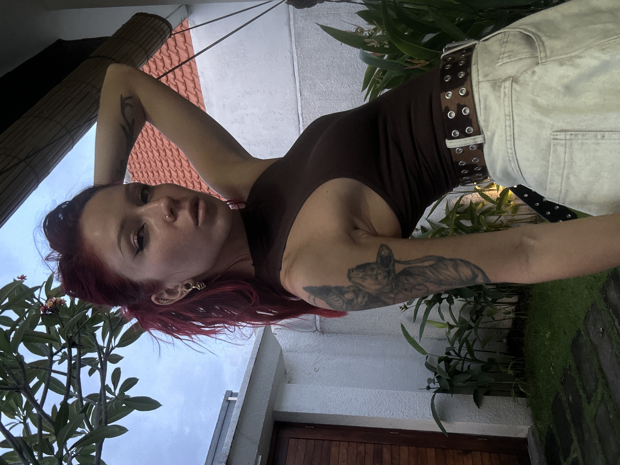 A medium shot captures a woman with striking red hair posing for a photo, seen from a low angle looking directly into the camera. She wears a dark brown sleeveless top and light-colored jeans, accessorized with a dark brown studded belt. Tattoos are visible on both arms, including a prominent animal figure on her right arm. She also has a nose ring and intricate gold earrings/cuffs on her left ear.
The setting is outdoors, likely a tropical environment in North Kuta, Indonesia. Above the woman, a wooden or bamboo roll-up blind or awning is visible, alongside a red-tiled roof and a white building wall. Lush green tropical foliage, including trees and smaller plants, frames the background, indicating a garden or courtyard area. Architectural elements like a white wall and a dark wooden door are also present in the lower part of the image, next to a patch of grass with stepping stones. The sky, visible in parts, suggests soft, diffused light, possibly indicating an overcast day or early evening. The overall mood is relaxed and natural. There is no visible text in the image.