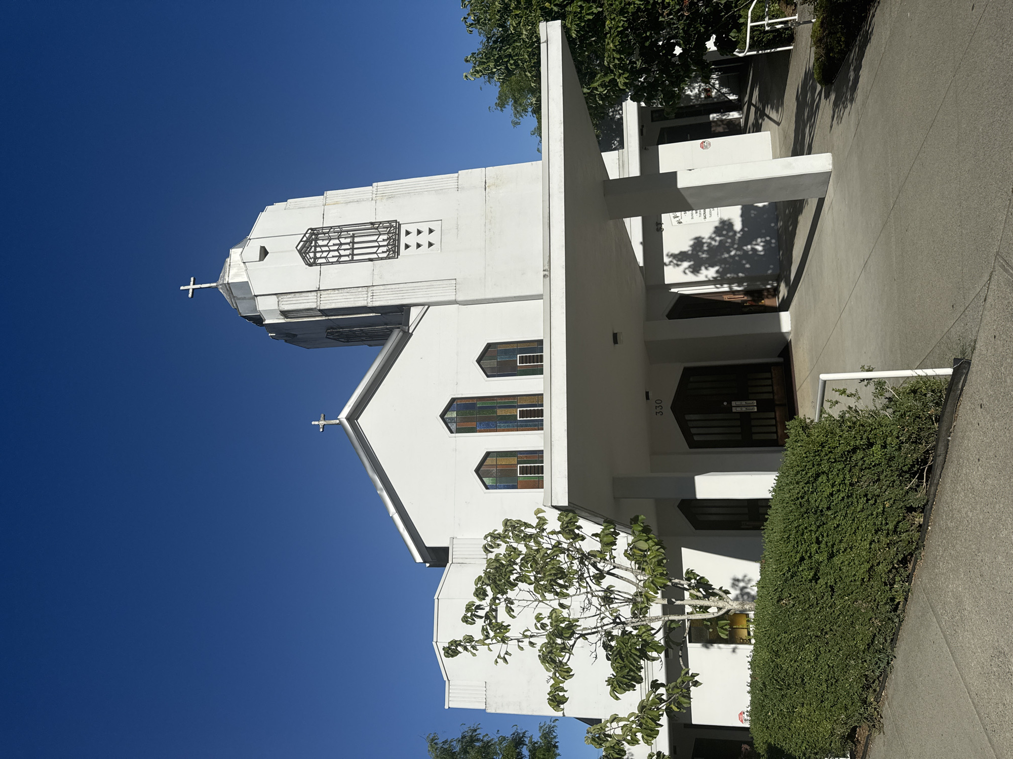 The image shows the exterior of a white church with a modern architectural style. A prominent feature is the tall, slender bell tower topped with a cross. The church has a large, covered entryway with columns supporting a flat roof. Stained glass windows, displaying a colorful geometric pattern, are visible on the facade. A neatly trimmed green hedge lines the walkway leading to the entrance. The sky is clear and blue. The church is located at 330 Agnes Street in the city of New Westminster, Canada. There are no people visible in the image.