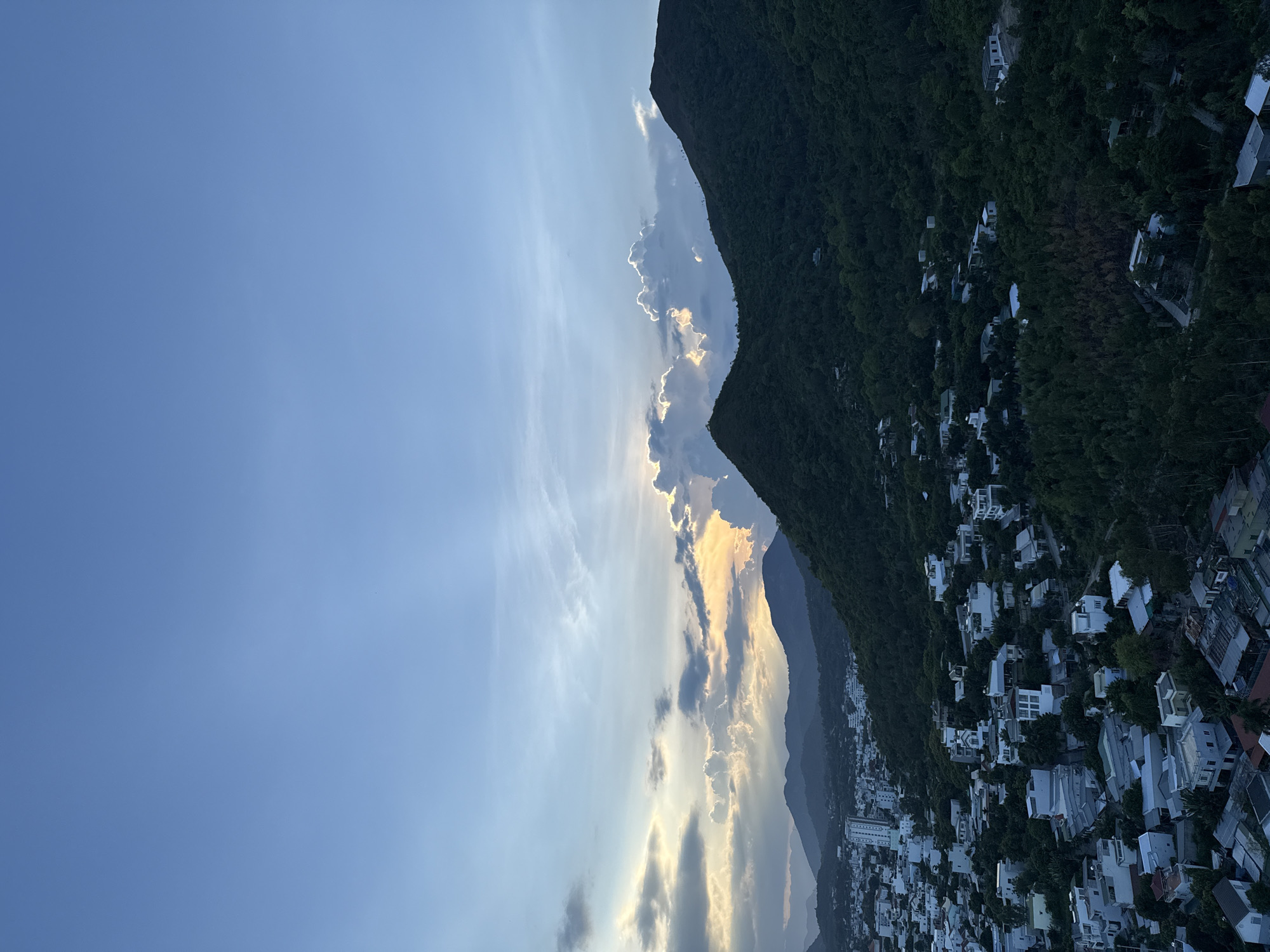 The image captures a panoramic view of Bac Nha Trang Ward, Vietnam, during what appears to be late afternoon or early evening. A vast, clear blue sky dominates the upper half of the frame, transitioning to a gradient of softer blues and grays towards the horizon. Wisps of white and gray clouds are scattered across the sky, with a particularly dramatic cluster illuminated by the setting sun, casting a warm, golden glow.
In the middle ground, lush green mountains rise, their slopes densely covered with trees. The texture of the foliage suggests a tropical environment. Nestled within the foothills and extending towards the foreground is a residential area. The buildings are predominantly white or light-colored, with varying roof types, indicating a mix of architectural styles. Houses are clustered together, interspersed with trees and vegetation, creating a sense of density. Some taller, modern buildings are visible among the lower-rise structures.
The lighting suggests a natural, diffused light source, likely the sun nearing the horizon, which creates subtle shadows and highlights on the landscape and buildings. The overall impression is one of a serene, developing urban landscape set against a dramatic natural backdrop. There are no visible people or immediate actions depicted in the scene. The focus is on the interplay of the urban environment, the natural topography, and the atmospheric conditions of the sky.