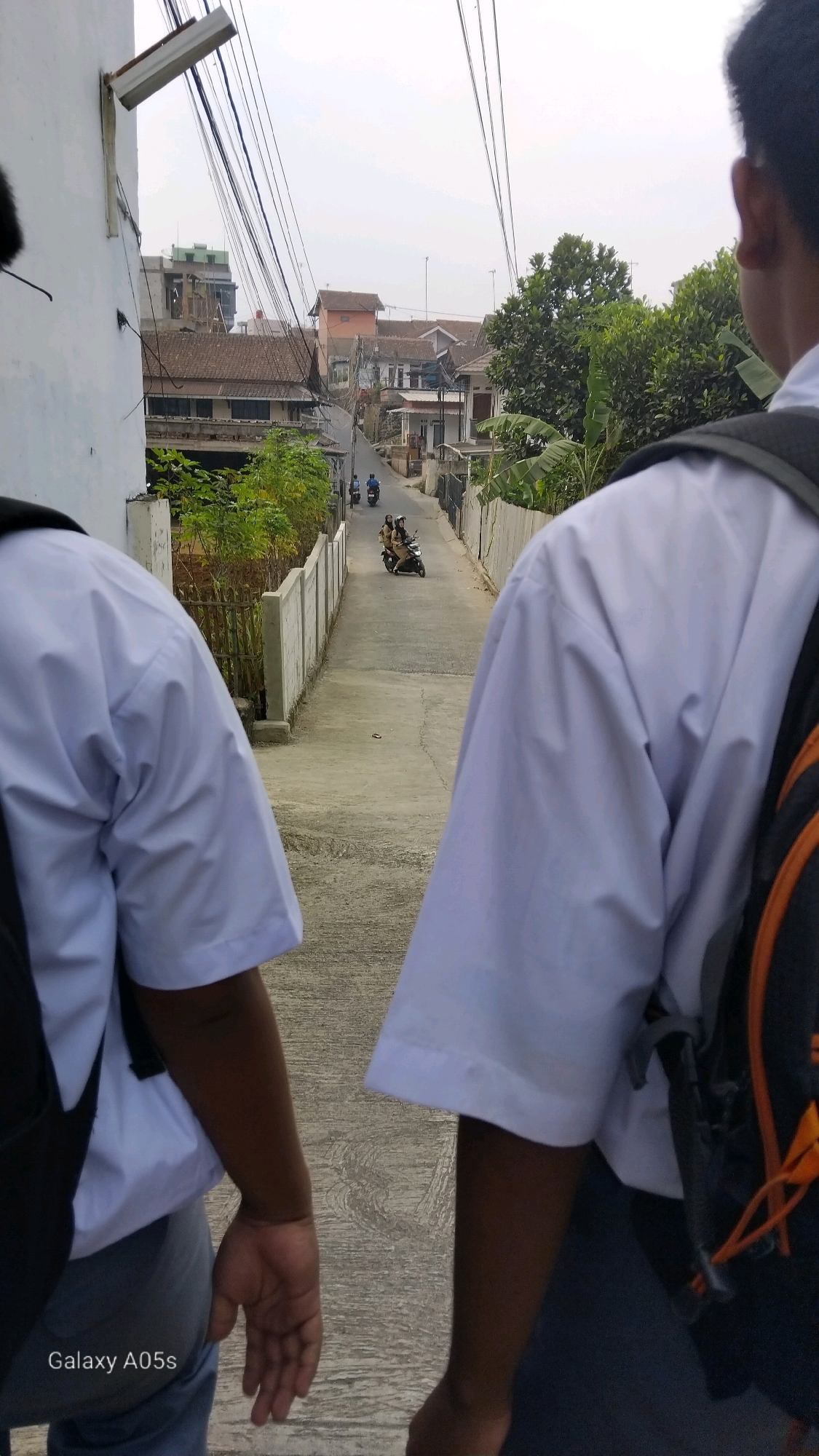Two individuals, appearing to be students in school uniforms, walk down a narrow, sloped street in Sumedang, Indonesia. They are viewed from behind, with their white shirts and backpacks dominating the foreground. The street is paved with concrete and is lined with a low concrete wall on the left and a more rustic fence on the right, partially obscured by green foliage.
Beyond the wall, residential buildings with tiled roofs are visible, interspersed with trees and utility poles carrying numerous electrical wires that crisscross the sky. In the distance, two motorcycles are traveling up the street. One motorcycle carries two passengers, a driver and a passenger, while a second, solitary motorcycle follows behind. The sky is overcast. The "Galaxy A05s" watermark is present in the lower left corner of the image, indicating the device used to capture the photograph.