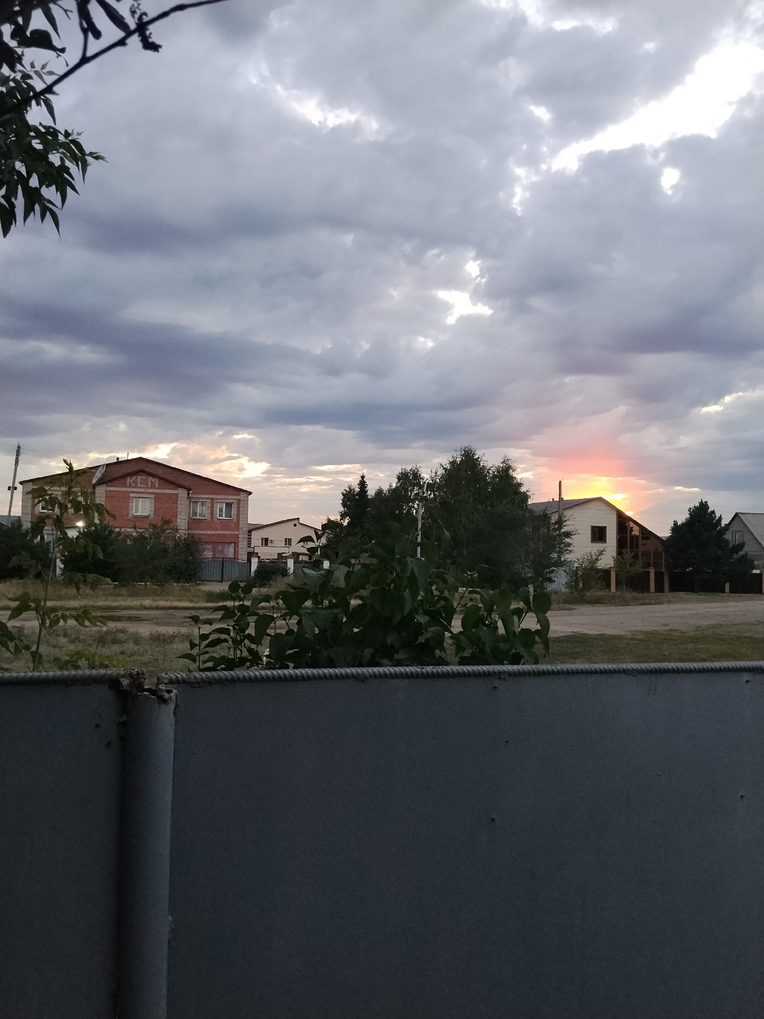 The image depicts a rural landscape under a cloudy sky, with a hint of sunset casting a pinkish-orange glow on the horizon. In the foreground, a dark metal fence with visible rope detailing runs across the bottom of the frame, partially obscuring a patch of dry grass and some leafy green bushes.
Behind the fence, several buildings are visible. The most prominent is a brick building with "KCM" written across its upper facade. To its right, a lighter-colored house with a dark roof is situated, with trees interspersed between the structures. The overall impression is one of a quiet, possibly semi-abandoned, semi-residential area. The presence of "KCM" on the building might suggest it's a commercial or industrial structure, possibly related to a local business or organization. Given the context of Kokshetau, Kazakhstan, this scene likely represents a suburban or outskirt area of the city, where industrial or commercial buildings might coexist with residential dwellings. The cloudy sky and fading light suggest it's either early morning or late evening. There are no people or animals visible in the frame.