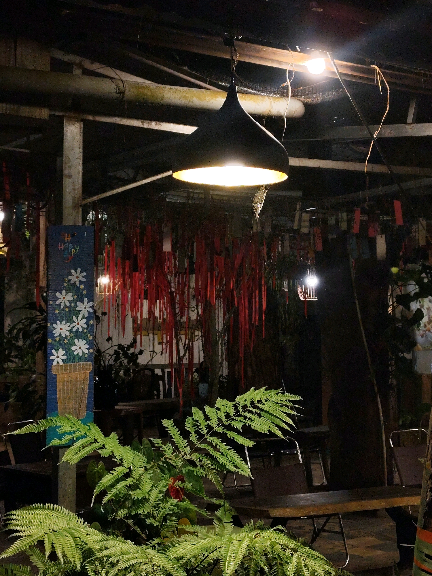 The image captures an outdoor seating area at night, likely a cafe or restaurant, in Da Lat, Vietnam. The scene is dimly lit by overhead lamps, one large black pendant light illuminating a portion of the foreground and several smaller, bare bulbs further back. The environment appears somewhat rustic and overgrown, with lush green ferns prominently featured in the foreground, obscuring a portion of the tables and chairs.
Behind the ferns, several wooden tables with metal-legged chairs are visible, set up in an open-air space. The background reveals a ceiling structure of exposed beams and pipes, some of which appear weathered or stained. Hanging from the ceiling are numerous thin, red streamers or ribbons, creating a decorative element. To the left, a tall, narrow blue panel with a hand-painted design of white flowers in a pot and the word "HAPPY" is mounted on a post. A dark tree trunk is visible towards the right side of the frame. The overall atmosphere suggests a relaxed, bohemian setting, possibly designed to blend with the natural surroundings of Da Lat, a city known for its cooler climate and abundant plant life.