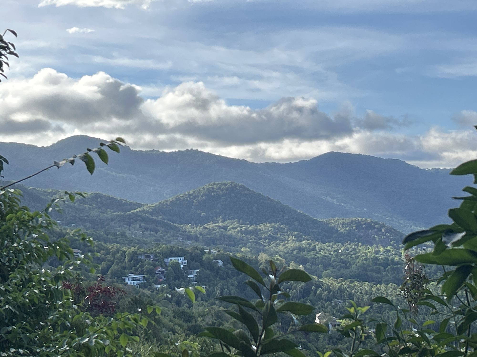 The image depicts a mountainous landscape in ตำบลเกาะพะงัน, Thailand, featuring a series of green, densely forested hills under a sky with scattered clouds. Several white structures, likely houses, are visible amid the vegetation on the lower slopes of the hills. The foreground includes foliage from trees and bushes, partially obscuring the view. The lighting suggests a daytime scene with slightly diffused sunlight.
