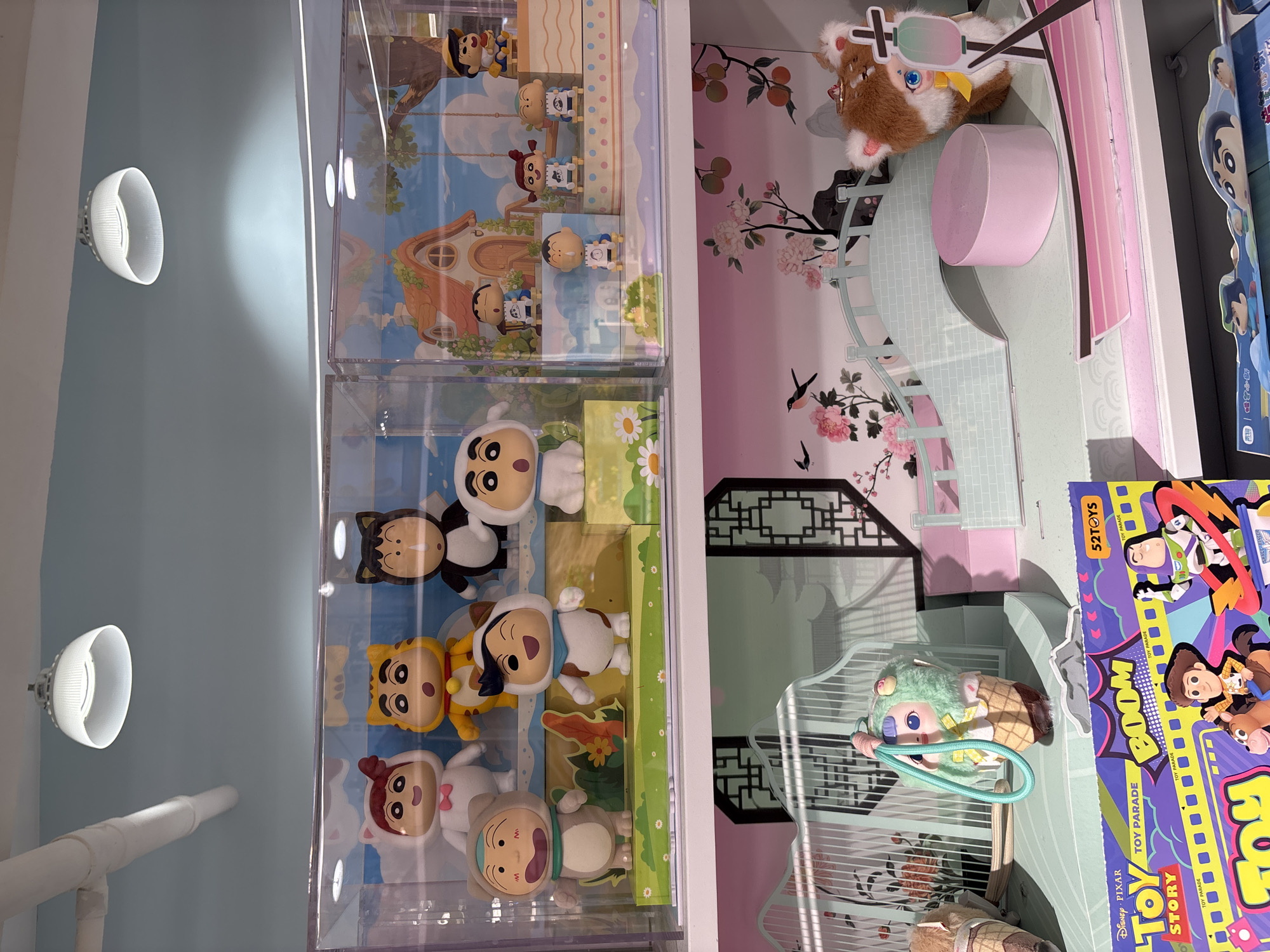 The photograph shows a display of toys, potentially located in a store or exhibition, possibly in the city of West Jakarta, Indonesia. The toys are contained within clear, possibly acrylic, display cases.
On the left side of the display, there are figures from the cartoon series "Pleasant Goat and Big Big Wolf". The figures are arranged on a background that includes grassy areas.
To the right of the "Pleasant Goat and Big Big Wolf" figures, there is a more elaborate display. It features a small plush toy that resembles a cat wearing sunglasses, along with other elements of a whimsical environment that include pink platforms, floral decorations, and what appears to be a decorative architectural structure. Adjacent to this arrangement is a box featuring characters from the Disney Pixar movie "Toy Story".
The background wall is light blue, and two circular lights are mounted on it. There is also a glimpse of white piping or a structural element against the blue background.