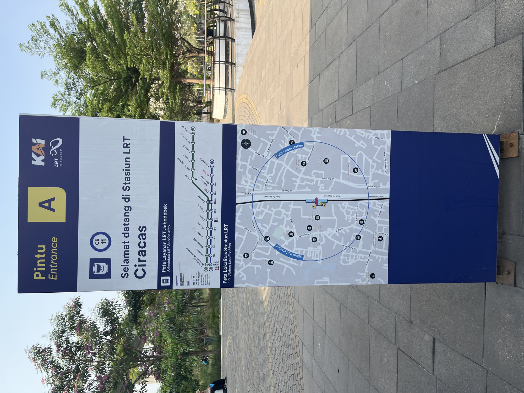 The image shows a tall, blue and yellow informational sign at an outdoor location in East Jakarta, Indonesia. The sign, which is positioned on a paved walkway, provides information about the Jabodebek LRT (Light Rail Transit) system. The top of the sign features the KAI LRT JABODEBEK logo and the text "Pintu Entrance A". Below this, there is a welcome message in Indonesian, "Selamat datang di Stasiun LRT Ciracas" (Welcome to Ciracas LRT Station). The sign includes a map of the Jabodebek LRT service lines and a locality map of the Ciracas LRT station, indicating nearby points of interest and a 500-meter radius from the station. In the background, there are trees, some with pink flowers, and a clear sky. Two people are visible in the background, one sitting on a bench and another walking. The ground is paved with large, grey square tiles.