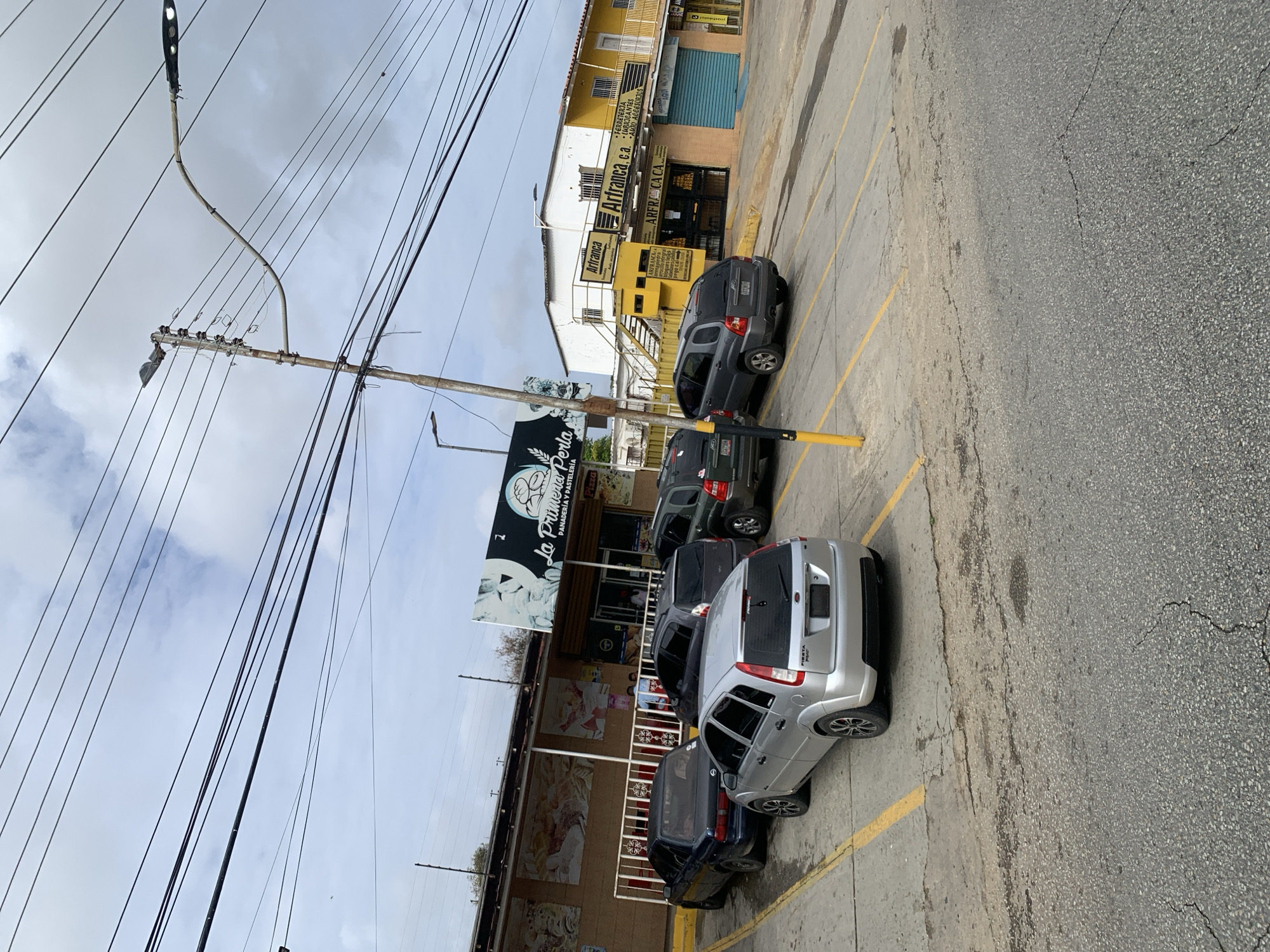 The image depicts a street scene in La Tagua, Venezuela, featuring a parking lot in front of commercial establishments. On the left, a building with a sign reading "La Primera Perla Panaderia y Pasteleria" is visible, with a mural on its facade. Several cars are parked in the lot, including a silver Ford Fiesta, a dark sedan, and two SUVs. In the background, a yellow building with signs for "Arfranca, c.a." and "Arfranca.c.a." is prominent. The environment includes utility poles with numerous overhead wires, a street lamp, and a cloudy sky. The ground surface is a mixture of cracked asphalt and concrete.