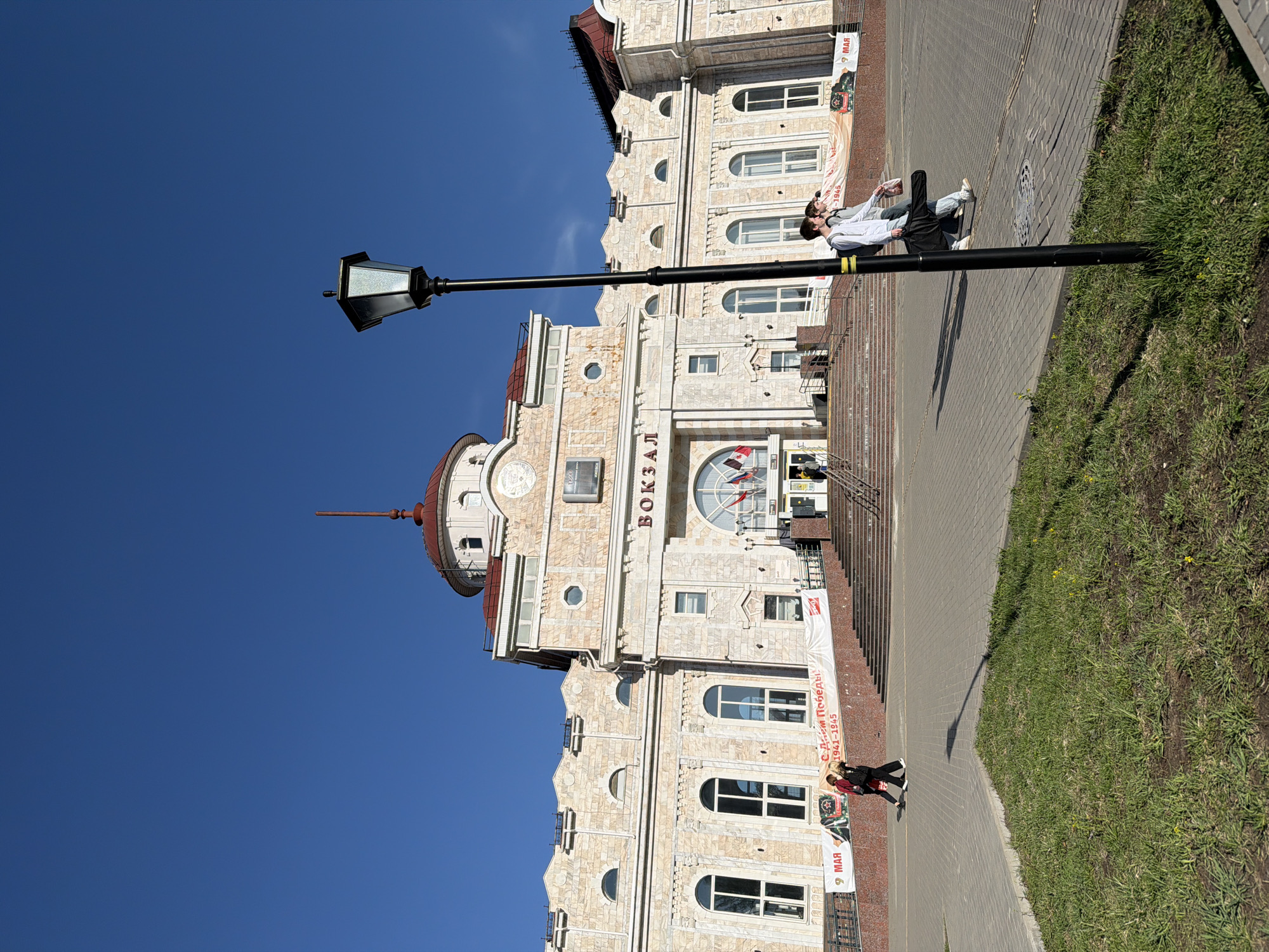 A street view in Izhevsk, Russia, presents a tall stone building labeled "вокзал" (Train Station), next to a black lamppost, under a clear blue sky. Two people are visible in the background near the steps, and another person is walking in the foreground next to the grassy area on the right side of the frame. Banners advertising the anniversary of Victory Day are hanging in front of the building.
