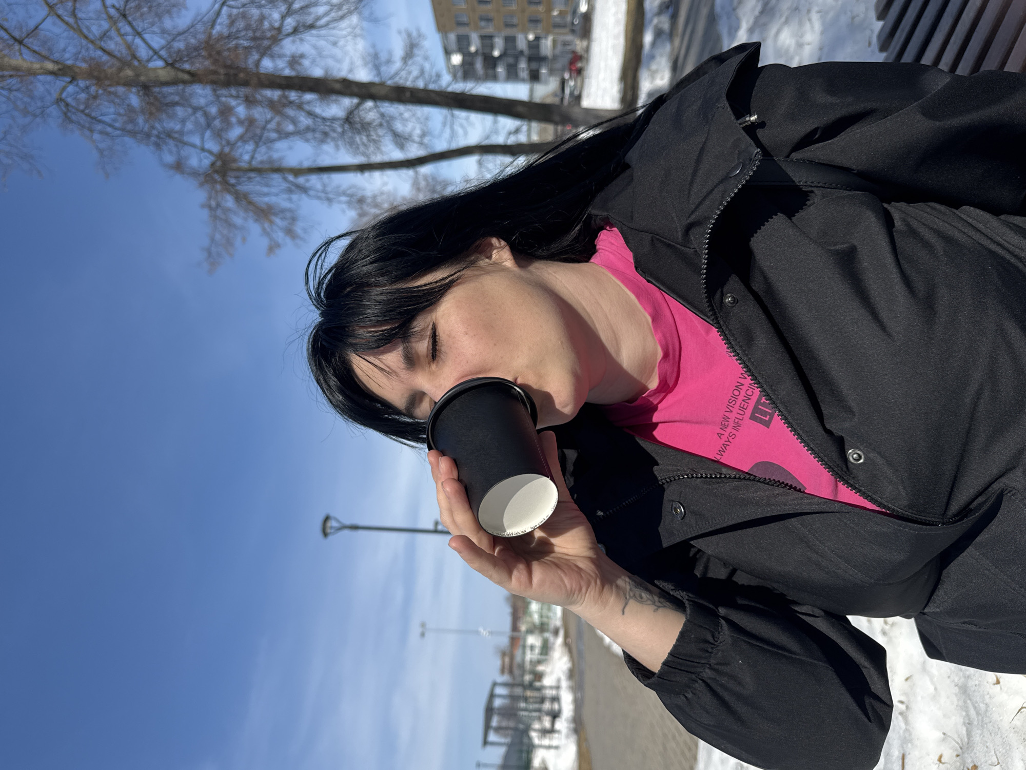 A woman with dark hair is drinking from a black coffee cup. She is wearing a pink t-shirt and a black jacket. The scene appears to be outdoors in Zav'yalovo, Russia, as evidenced by the snowy ground, trees, street lamps, and buildings in the background.
