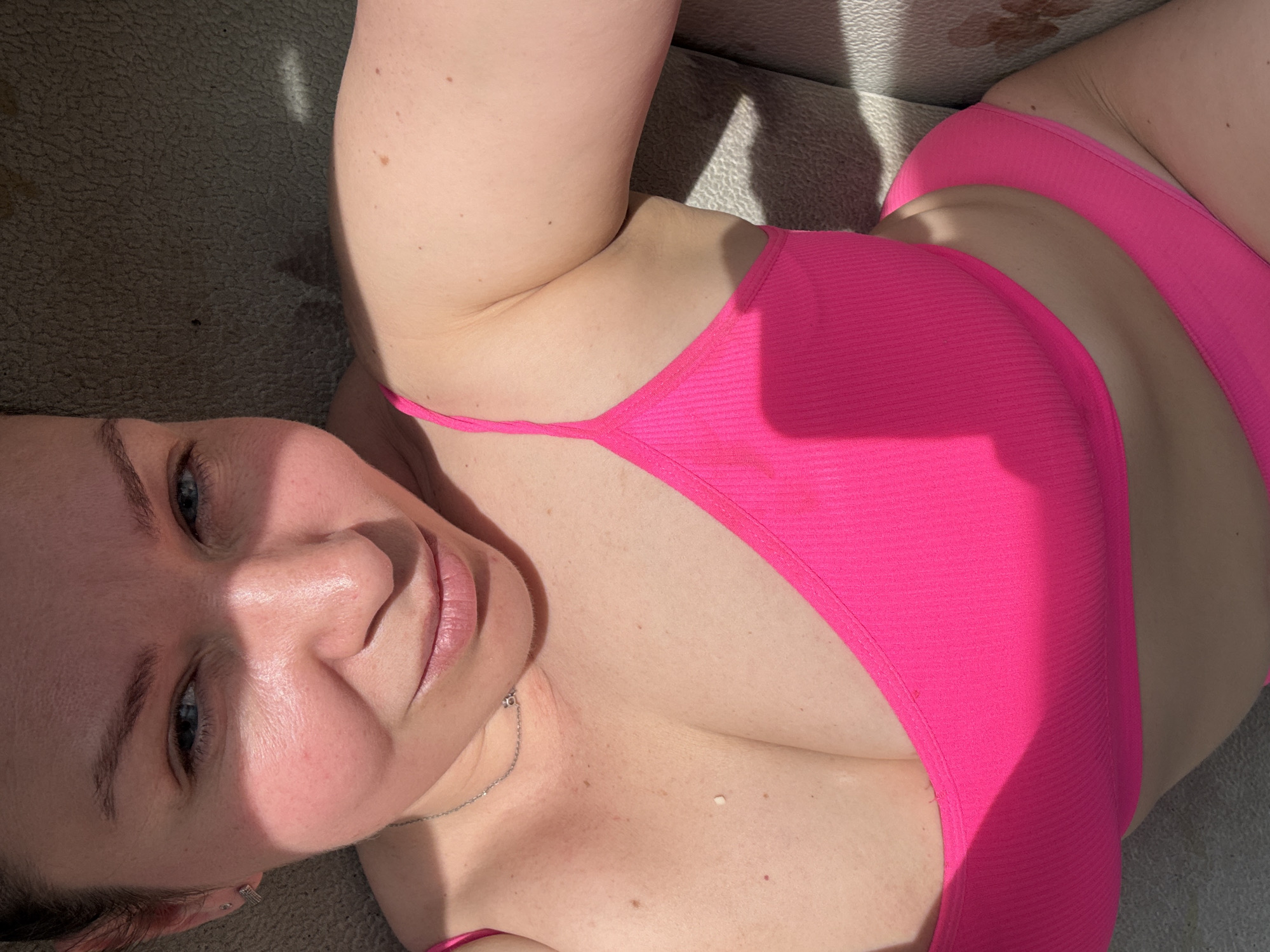 A fair-skinned woman lies in a pink bikini. She is indoors, possibly on a light gray textured surface. Part of her face is in shadow from a light source above. She is wearing a delicate silver necklace. The location is unknown and cannot be determined to be Chaikovsky, Russia from the given image.