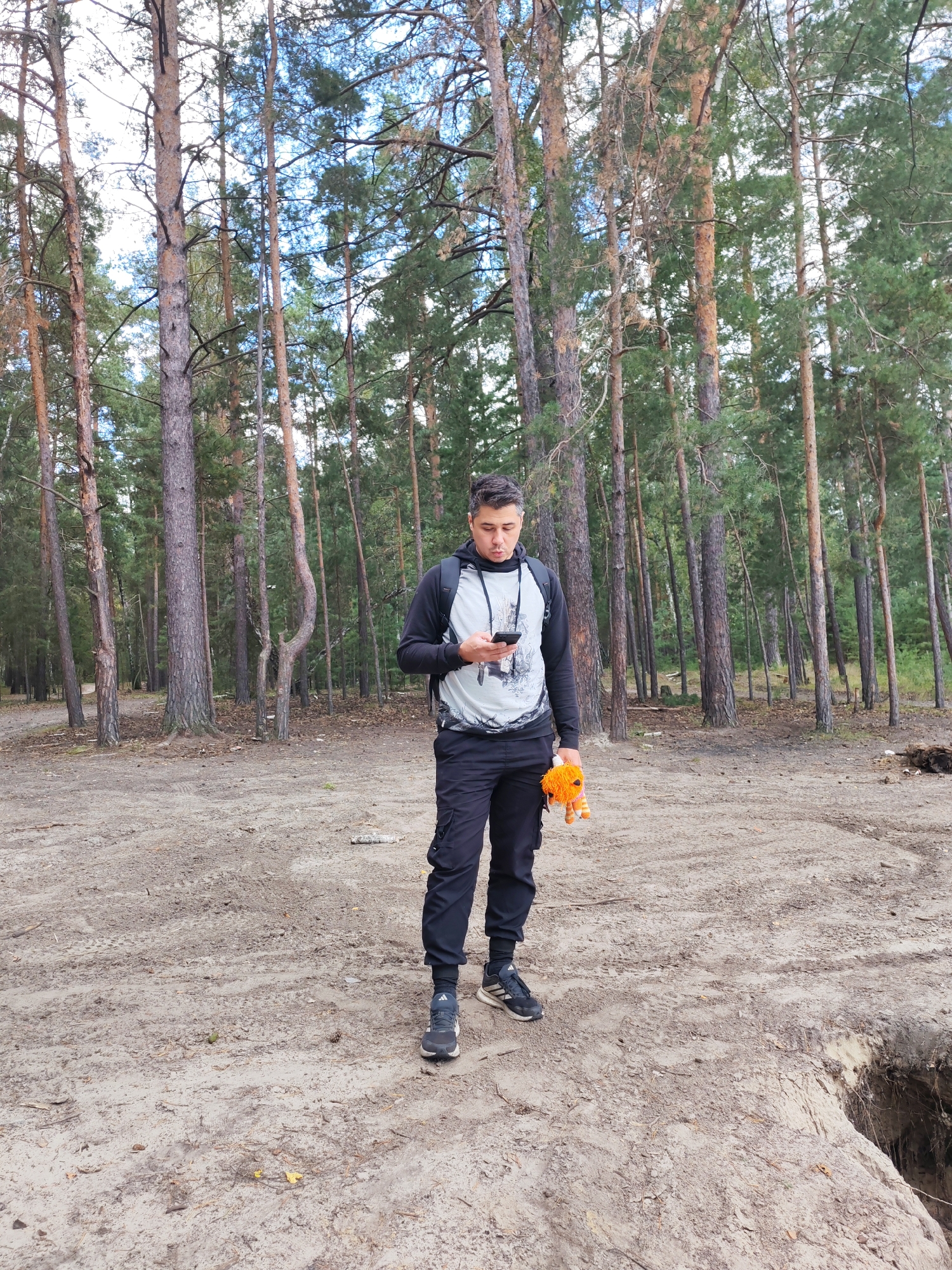 A young man stands in a forest clearing, looking at his smartphone. He is wearing a black hoodie with a gray design on the front, black cargo pants, and dark athletic shoes. He is holding a small, orange stuffed toy in his right hand. The ground is sandy and appears to be a natural path or clearing in a pine forest. There is a deep hole or ditch on the right side of the frame, possibly a sinkhole or excavation. The trees are tall and slender, with some dry branches visible. The sky is overcast. The image does not contain any specific visual cues to identify the location as Kurgan, Russia.