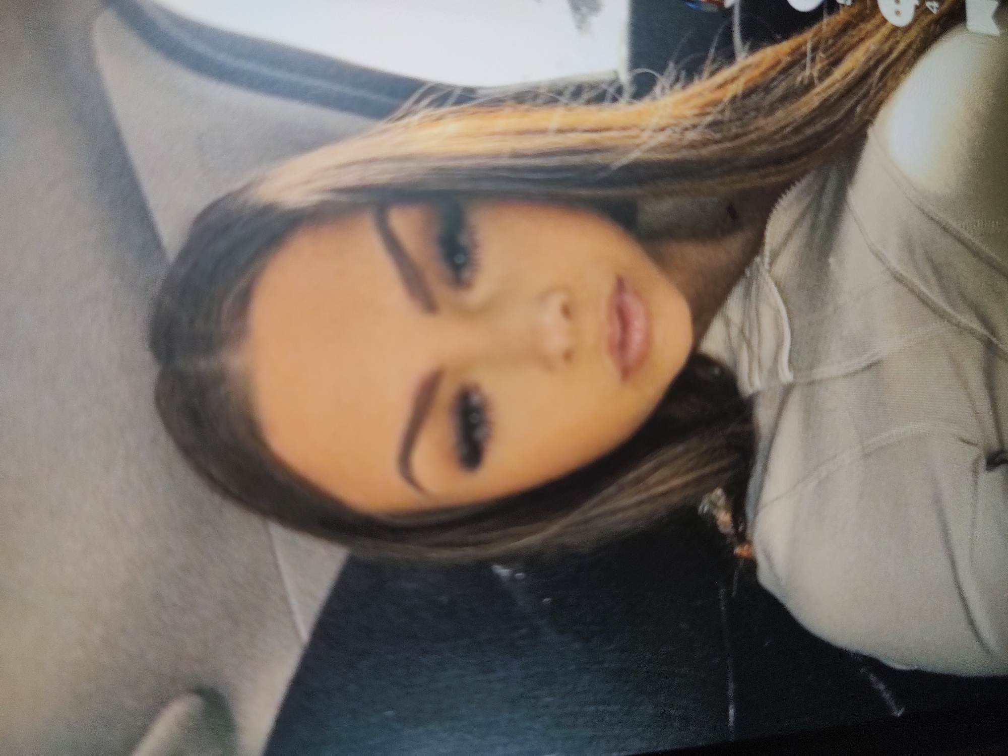 A woman with brown and blonde hair, heavy makeup including false eyelashes and contoured eyebrows, is wearing a light-colored zip-up jacket. The background appears to be the interior of a car, with a gray headrest visible on the left. The location is unknown.