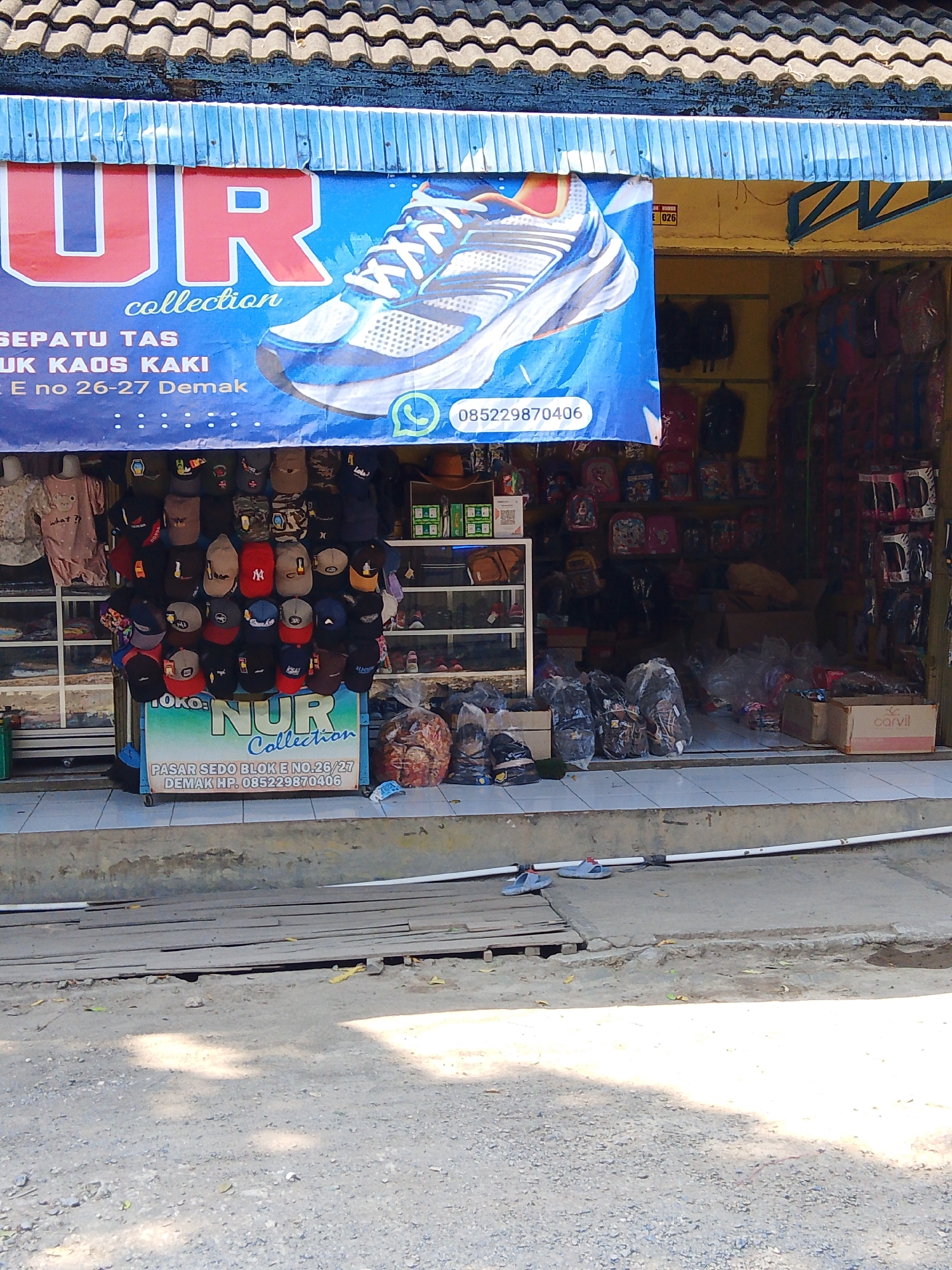 The image depicts a street-level view of a small retail establishment in Kedondong, Indonesia, identified by the signage "NUR Collection." The storefront is adorned with a blue banner featuring a large image of a sneaker, the store name, and contact information. The store appears to specialize in selling shoes, bags, and possibly apparel, as evidenced by the display of various hats, backpacks, and clothing items within and just outside the shop. Several plastic bags containing merchandise are also visible. The storefront has a tiled facade and is situated on a concrete platform. The street in front of the store is unpaved, with loose gravel and some wooden planks. Two pairs of sandals are placed on the concrete in the foreground, suggesting recent activity or display. The overall impression is that of a modest, local business operating in a semi-urban or rural setting.