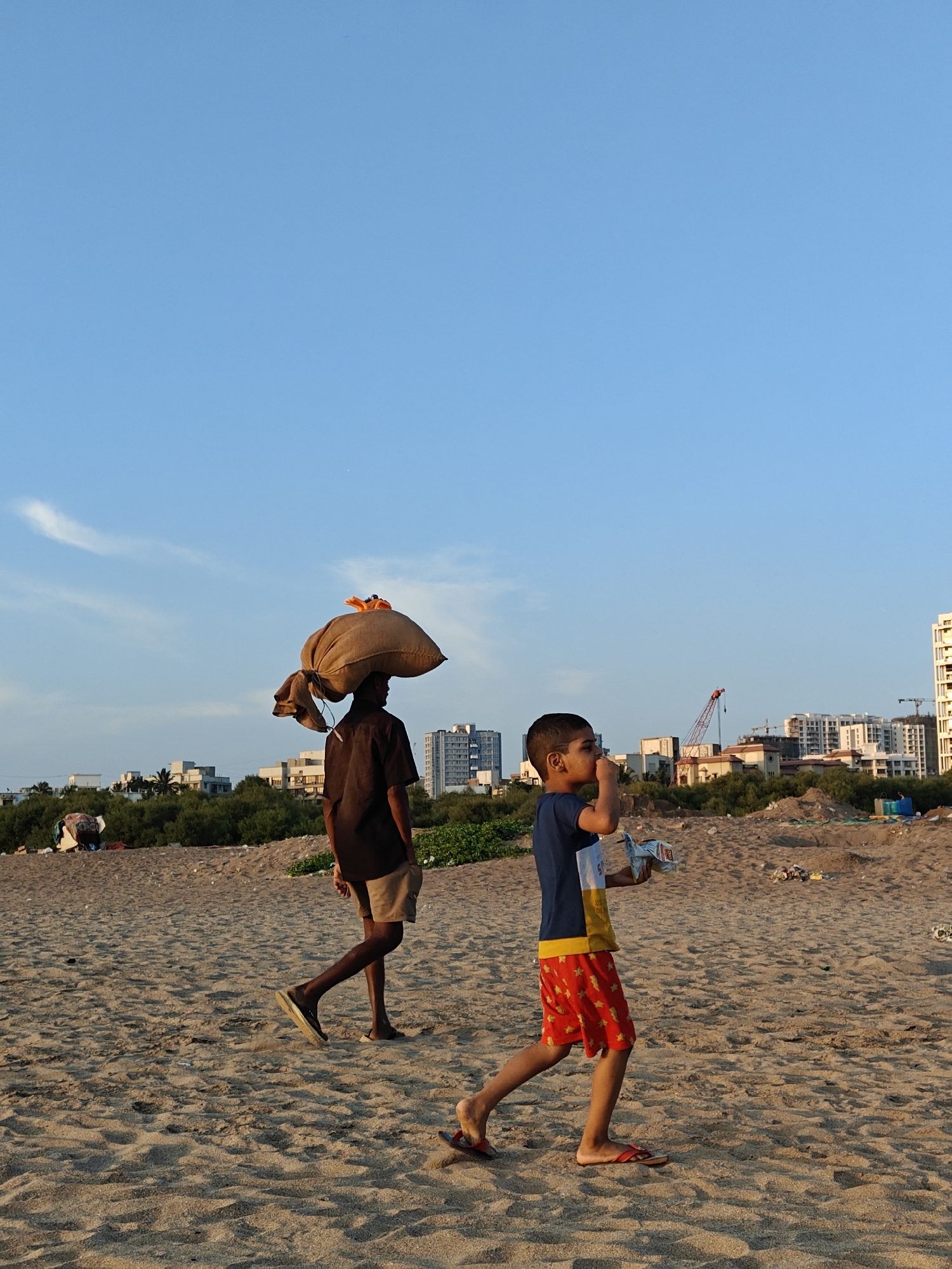Two boys walk across a sandy beach. The boy in the foreground holds a small white container to his mouth. He wears a short-sleeved blue and yellow shirt and red shorts with a yellow pattern, and red flip-flops. Behind him, a person carries a large sack on their head, wearing a dark short-sleeved shirt and khaki shorts. The person also wears dark sandals. In the background, buildings and a construction crane are visible against a blue sky. The location is likely a beach in Mumbai, India. The sand appears to have debris.