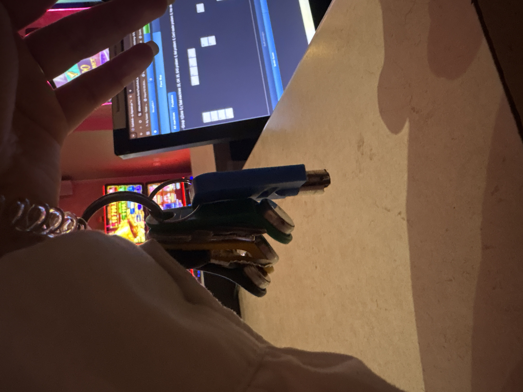 The image shows a close-up view of a hand holding a set of keys with various colored key fobs, including blue, green, and yellow. The keys are attached to a coiled metal keychain. In the background, out of focus, are elements that suggest a casino or gaming environment. There are brightly lit slot machines with colorful displays and a computer monitor showing a grid of information, possibly related to machine status or sales data. The scene appears to be indoors, with a tan-colored countertop in the foreground casting a shadow. The specific location in the city of Karlovo, Bulgaria, cannot be determined from the visual information provided in the image.