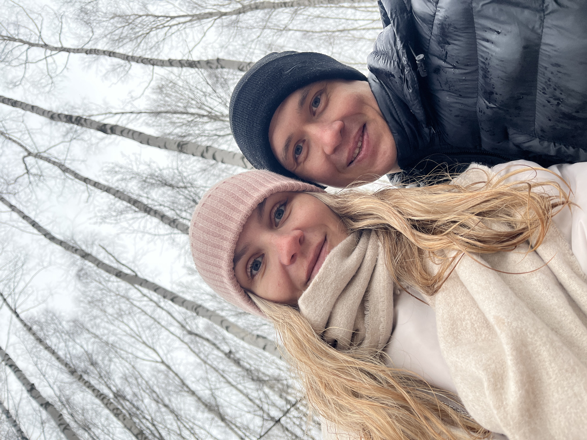 A selfie-style photograph captures two people outdoors, with a forest of birch trees in the background. The trees are leafless, suggesting a winter or late autumn setting. Overcast skies create diffused lighting.
The two individuals are positioned closely together. The person on the left has blonde hair, styled long and loose, and is wearing a pink knit hat. A light-colored scarf is wrapped around their neck. The person on the right is wearing a black knit hat and a black puffy jacket.
The photo's tilt and perspective imply the image was taken at an angle. The specific location is the city of Chaikovsky, Russia.