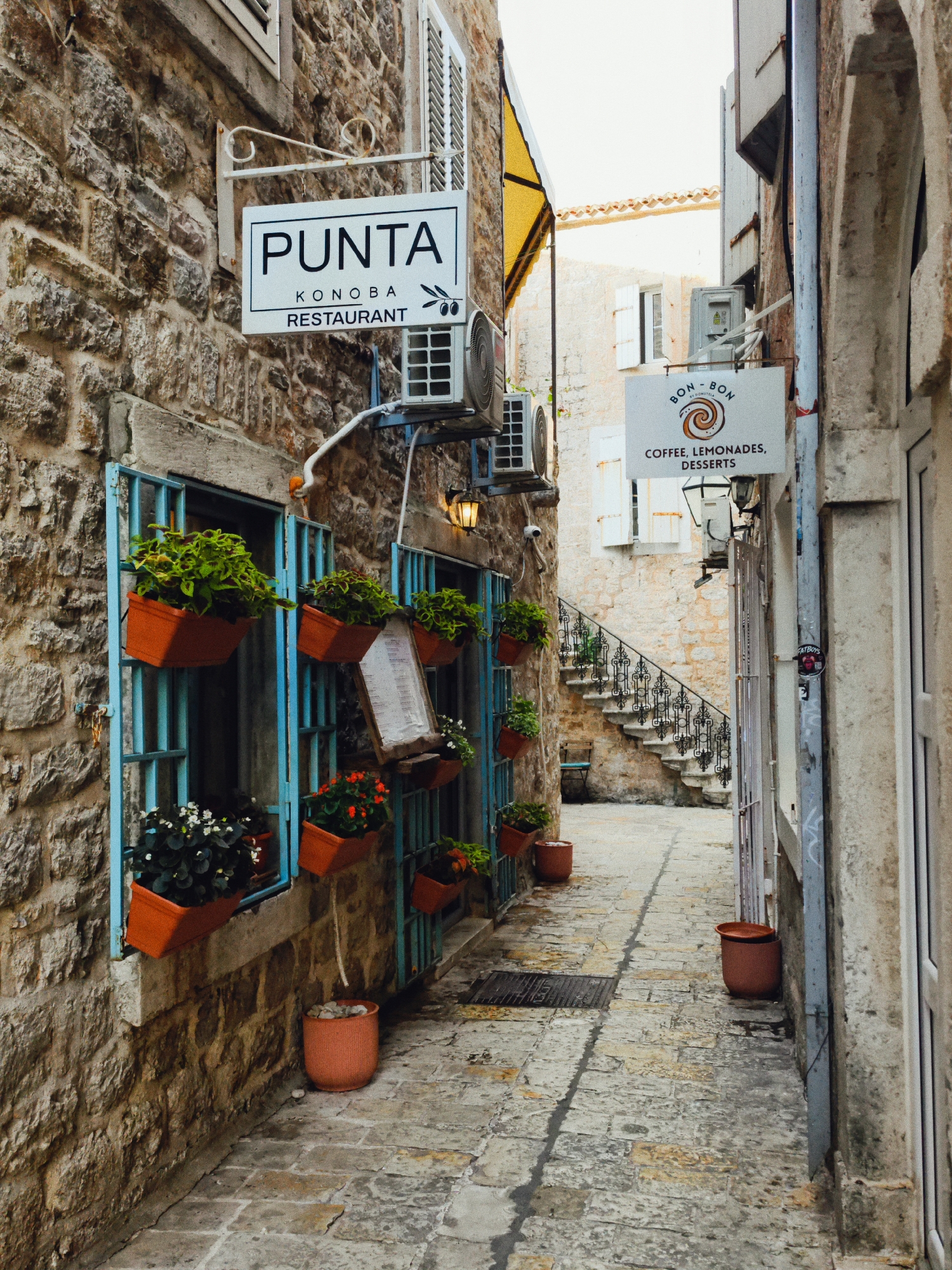 The image shows a narrow cobblestone alleyway in the Old Town of Budva, Montenegro. On the left, a weathered stone building has an open window with blue metal bars, adorned with several orange planters containing green and white flowers. A sign above the window reads "PUNTA KONDBA RESTAURANT". On the right side of the alley, another building has a sign that says "BON - BON COFFEE, LEMONADES, DESSERTS". Further down the alley, a stone staircase with an ornate metal railing leads upwards. The alley is sparsely decorated with potted plants and a few terracotta pots. The overall impression is that of a quaint and historic European street.