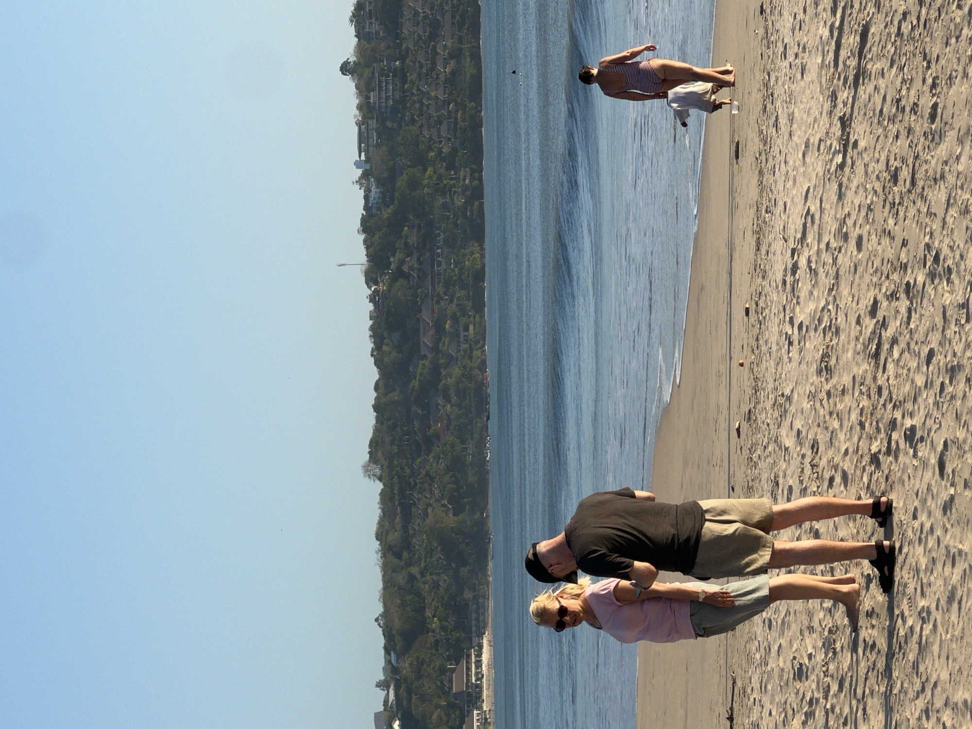 The image depicts a sandy beach in Kuta Selatan, Indonesia, with the ocean and a treed hillside in the background. On the beach, a couple stands facing each other in the foreground. The woman, with blonde hair, wears sunglasses and a pink t-shirt with denim shorts. The man, wearing a black t-shirt and khaki shorts, has a black baseball cap on and is looking at his phone. Further down the beach, a woman in a striped swimsuit walks towards the water, and a child in a white garment is crouching near the water's edge. The sea exhibits gentle waves, and the sky is clear and blue. The hillside behind the beach is covered with lush green trees and features buildings with red roofs and white facades, suggesting a resort area.