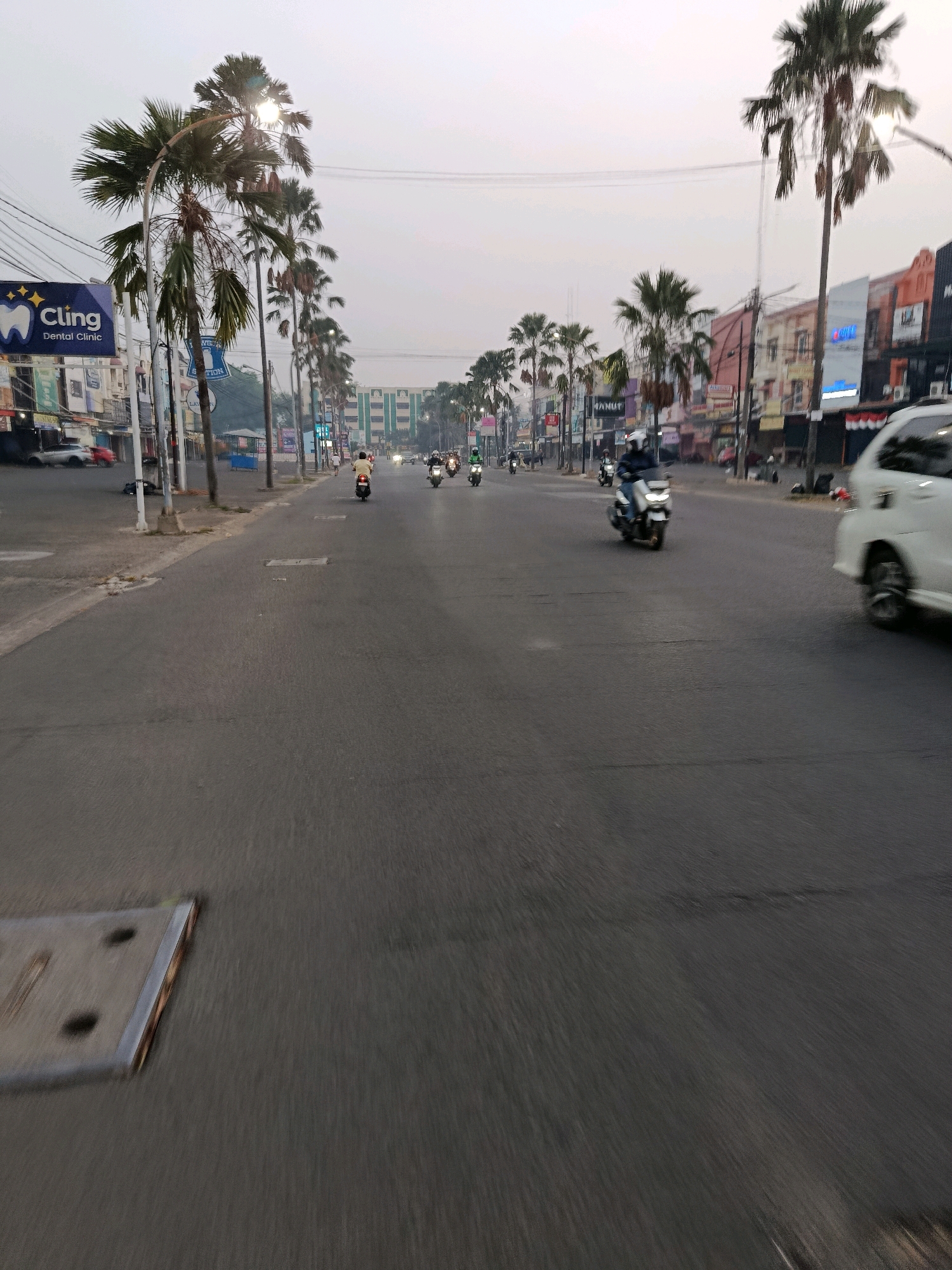 This is a medium-angle, forward-facing, shot of a street in the city of Bekasi, Indonesia. Palm trees line the left side of the street, and buildings with signs are visible in the background. Several motorcycles are on the road, with riders wearing helmets. A white car is on the right side of the road. The sky is overcast. The image appears to have been taken at dusk or dawn, due to the low light conditions. There is a manhole cover on the lower left of the image.