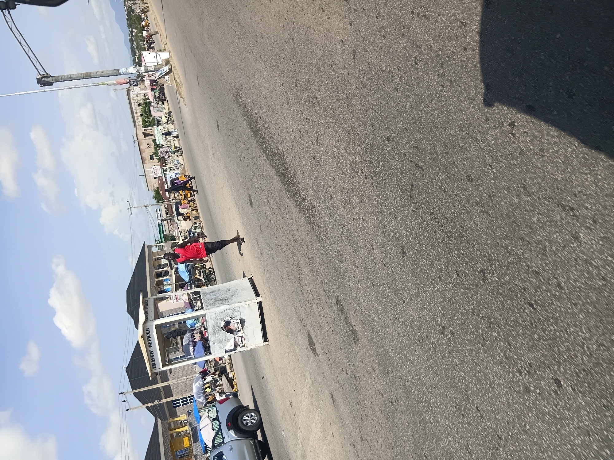 The image depicts a street scene in Jalingo, Nigeria, likely on a sunny day with scattered clouds. In the foreground, a man in a red tank top and dark pants walks away from a traffic officer booth. The booth is a light-colored, hexagonal structure with "TRAFFIC OFFICER" written on it, and several umbrellas are set up around it, suggesting a marketplace or roadside stalls. To the left of the booth, a silver pickup truck is parked. In the middle ground, various people and vehicles, including what appears to be a yellow tricycle, are visible on the road and sidewalk. Buildings with dark roofs line the background, and further in the distance, hills or mountains are discernible. The overall impression is of a busy, urban environment.