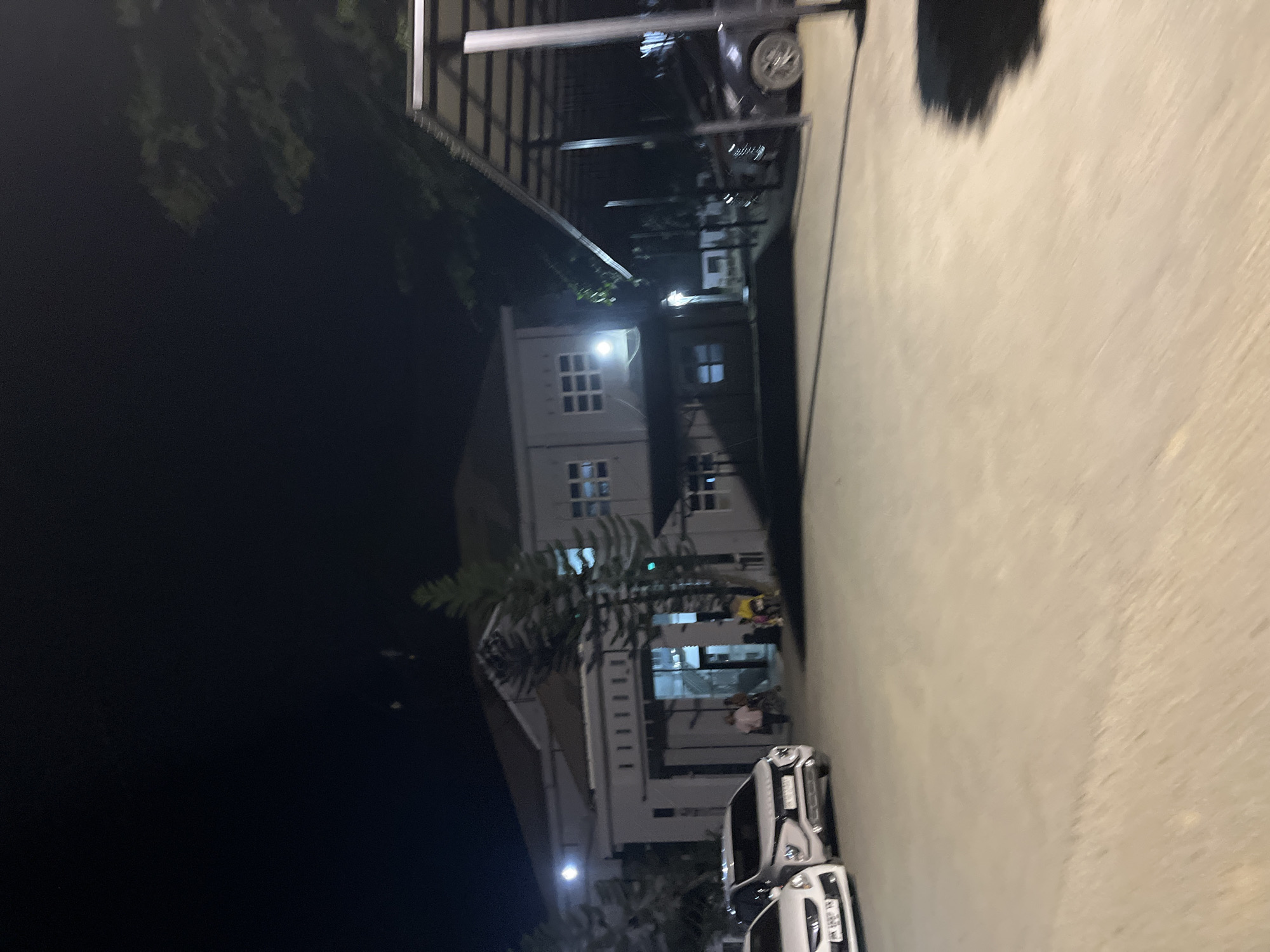 The image captures a nighttime scene in what appears to be a residential or commercial area in Kesehatanbandar, Indonesia. The orientation of the image is skewed, making it challenging to fully discern details.
In the foreground, two white vehicles are parked on a paved surface. Their headlights are not illuminated, suggesting they are not in active use. Further into the scene, a multi-story building with a modern architectural style is visible. The building is predominantly white and features numerous windows with a grid-like pattern. Some of these windows are illuminated, indicating interior lights are on.
A palm tree, its fronds dark against the night sky, stands between the viewer and the building, adding a touch of tropical greenery to the urban landscape. Above and behind the building, a darker structure with a slatted roof is visible, possibly a carport or an extension of the building. A car is partially visible under this structure.
The overall lighting suggests it is nighttime. The atmosphere appears calm and quiet. There are no obvious signs of any particular event or activity taking place, other than the presence of vehicles and illuminated buildings. The environment suggests a typical urban or suburban setting at night. No visible text is present in the image.