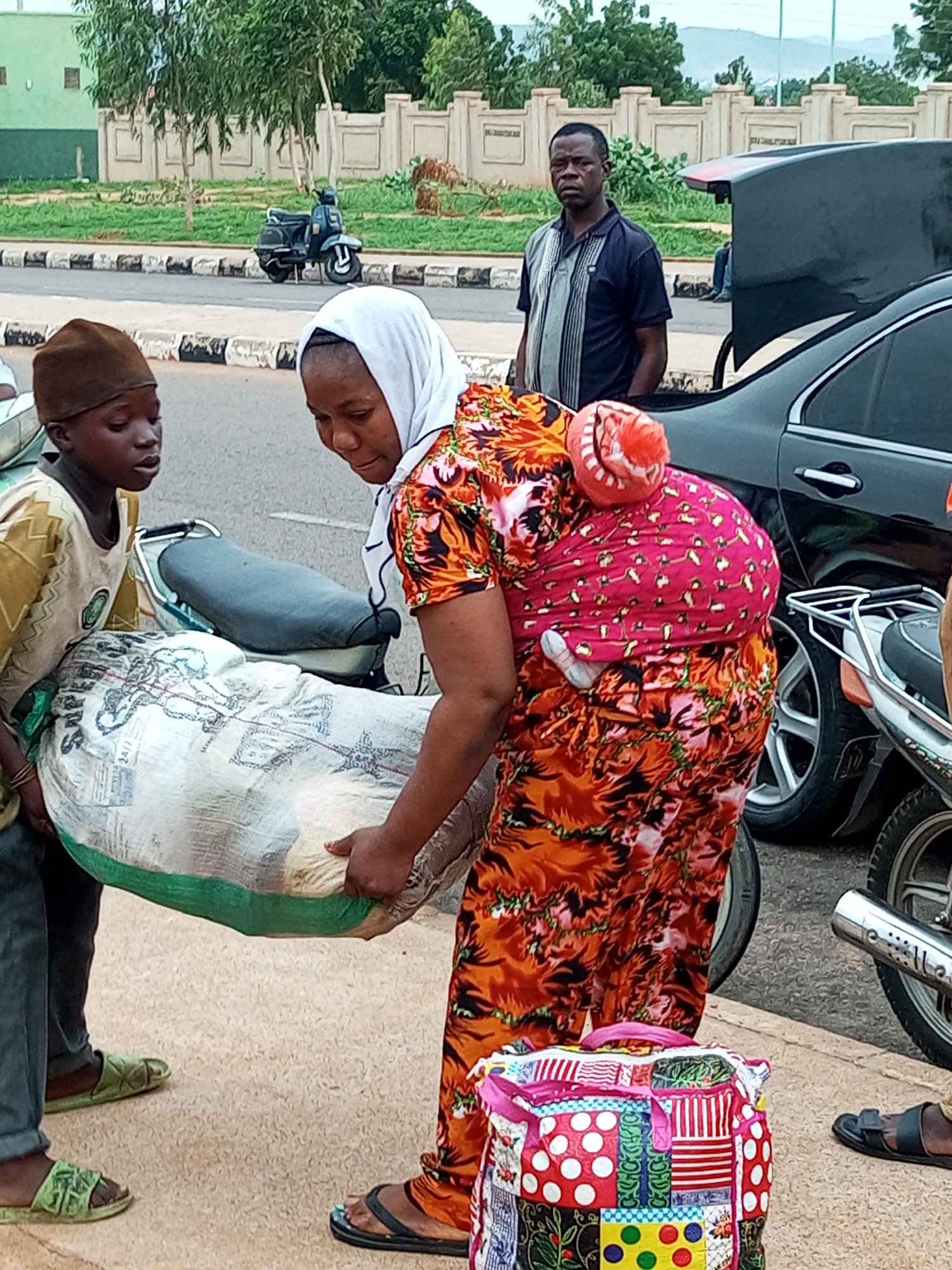 In the city of Gombe, Nigeria, on what appears to be a paved road with a sidewalk, a young boy wearing a brown cap and a light-colored shirt is assisting a woman. The woman, dressed in a vibrant orange and red patterned dress and a white headscarf, is carrying a large, light-colored bag wrapped in green material. She is using a baby carrier to support a child on her back, the child's head visible and wearing an orange and white striped hat. Beside them, a black car with its trunk open is parked, and a man in a blue and gray striped polo shirt stands near it, looking towards the camera. In the background, a dark-colored scooter is parked, and a low beige wall with a green-roofed building and trees can be seen beyond the road, with distant hills under a hazy sky. A colorful, patchwork bag sits on the ground near the woman's feet.