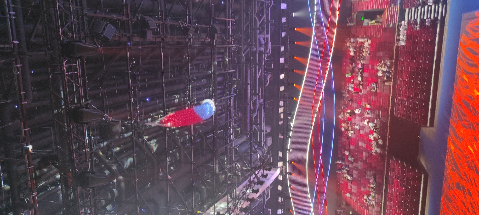 The image shows an indoor arena with rows of red seats filled with spectators. A large, inflatable object, red and blue, is suspended from the ceiling. The ceiling structure is elaborate, with a complex network of beams, pipes, and lighting equipment. The lower part of the image displays a stage area with an abstract, fiery pattern. The scene is likely a performance or sporting event. There is no information in the image to suggest the location is Moscow, Russia.