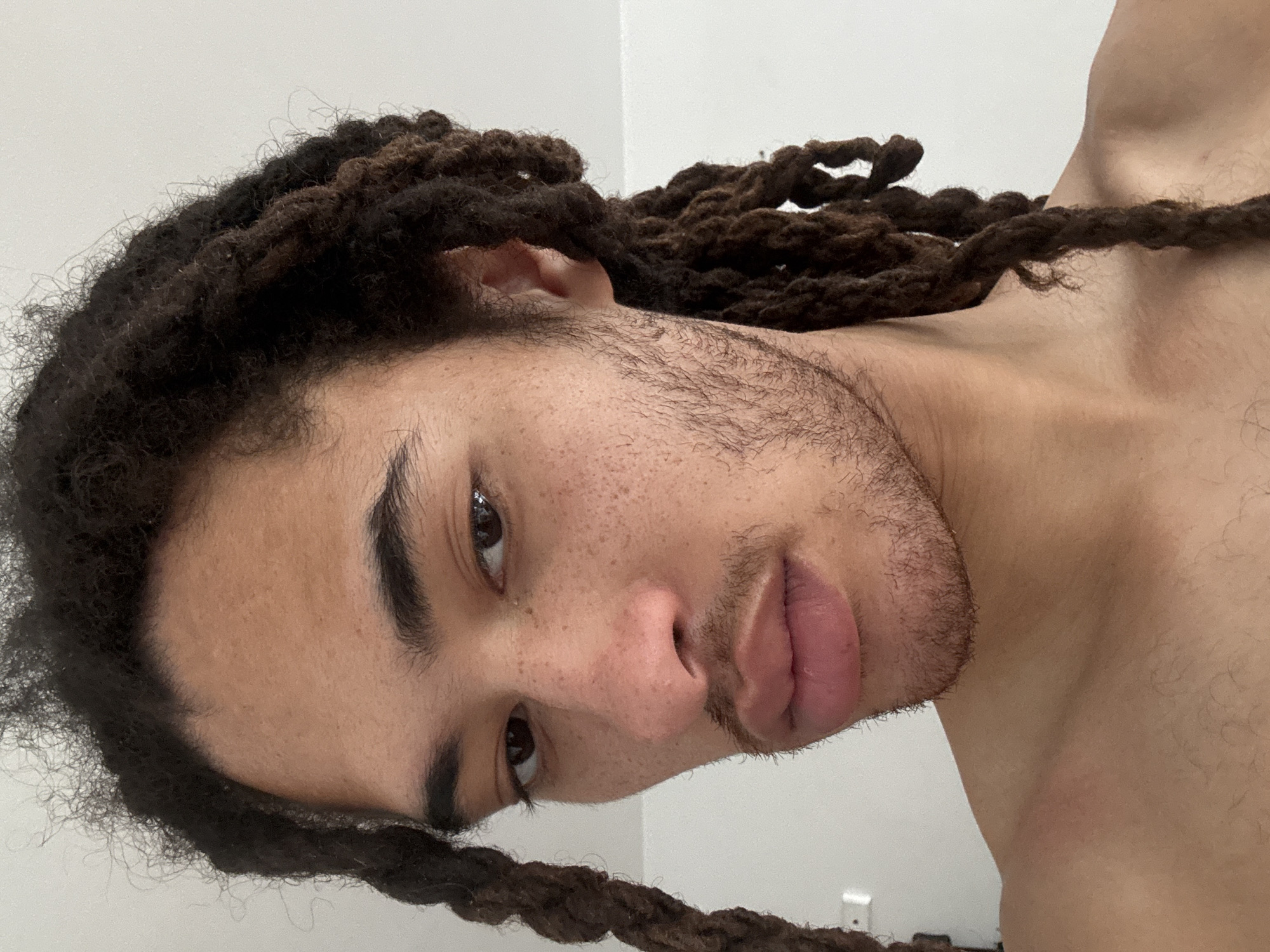 A close-up, angled portrait of a fair-skinned individual with dark brown hair styled in dreadlocks. Their face features dark eyes, prominent eyebrows, a slightly parted mouth with pink lips, and visible freckles on their nose and cheek. They have stubble or a light beard. The background appears to be a plain, light-colored wall. The individual is topless. The image does not appear to be in Paris, France. There is no indication of a location.