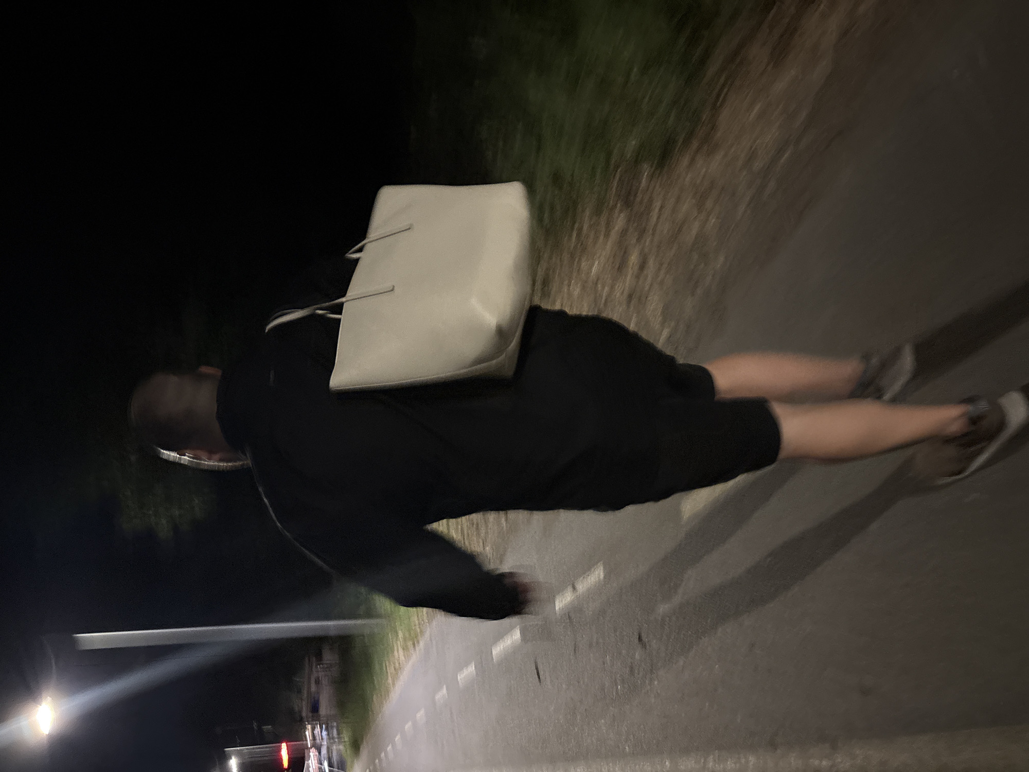 A person, seen from behind and in motion, walks on a paved path at night. They are wearing black shorts and a black shirt and carrying a large, light-colored tote bag over their right shoulder. The path has white dashed lines indicating lanes. A street lamp illuminates the scene from the left, casting long shadows. In the background, there are trees and what appears to be vegetation. The overall environment suggests an outdoor urban setting, possibly a park or a pathway along a road in Burgas, Bulgaria.
