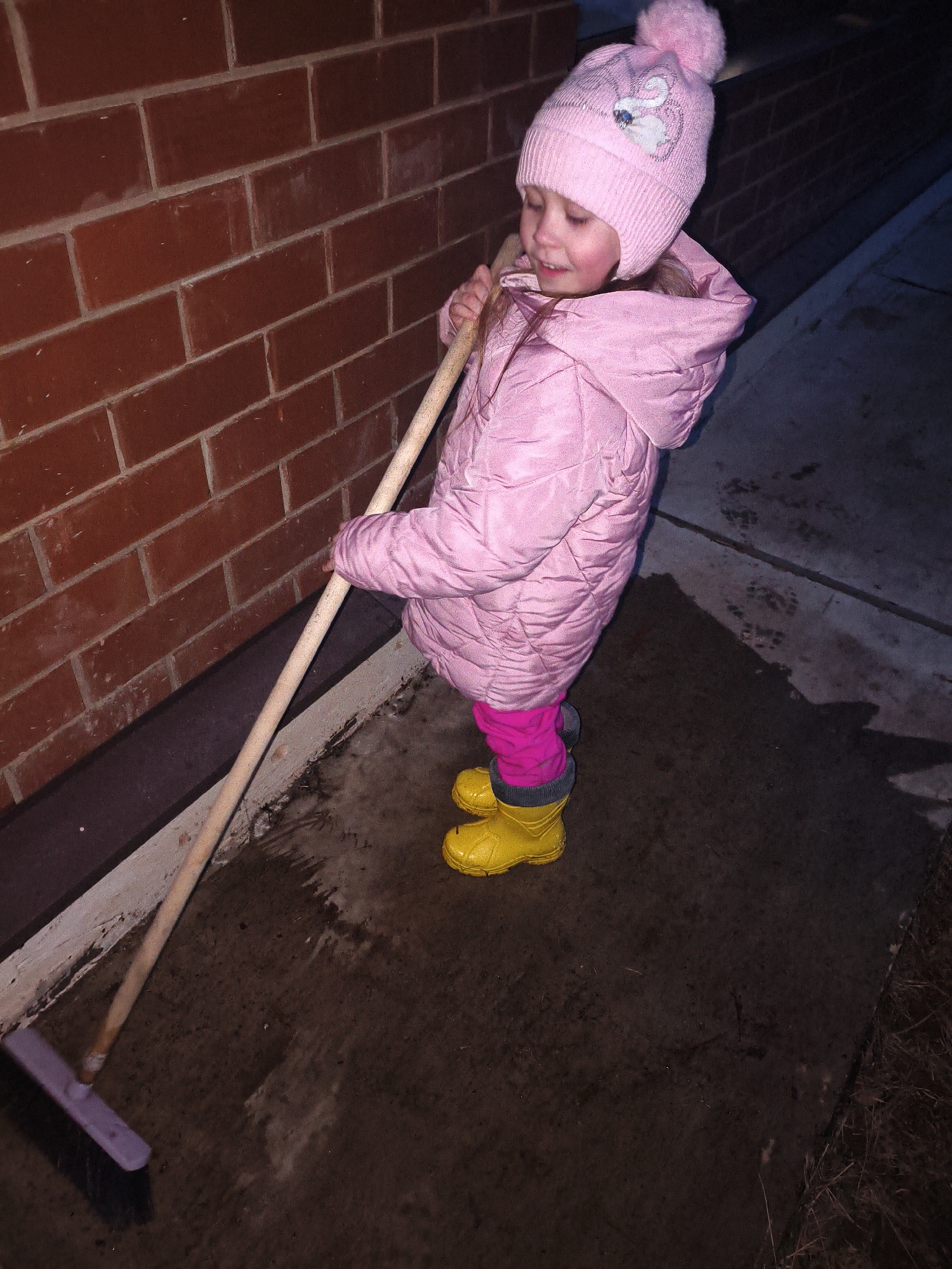 A young child in a pink winter coat, pink hat, pink leggings, and yellow rubber boots is standing outside next to a red brick wall. The child is holding a wooden broom with a purple head and appears to be sweeping a wet, dark grey pavement. The setting appears to be outside in a dim or shadowy area with a darker area of pavement stretching away. The location is Chaikovsky, Russia.