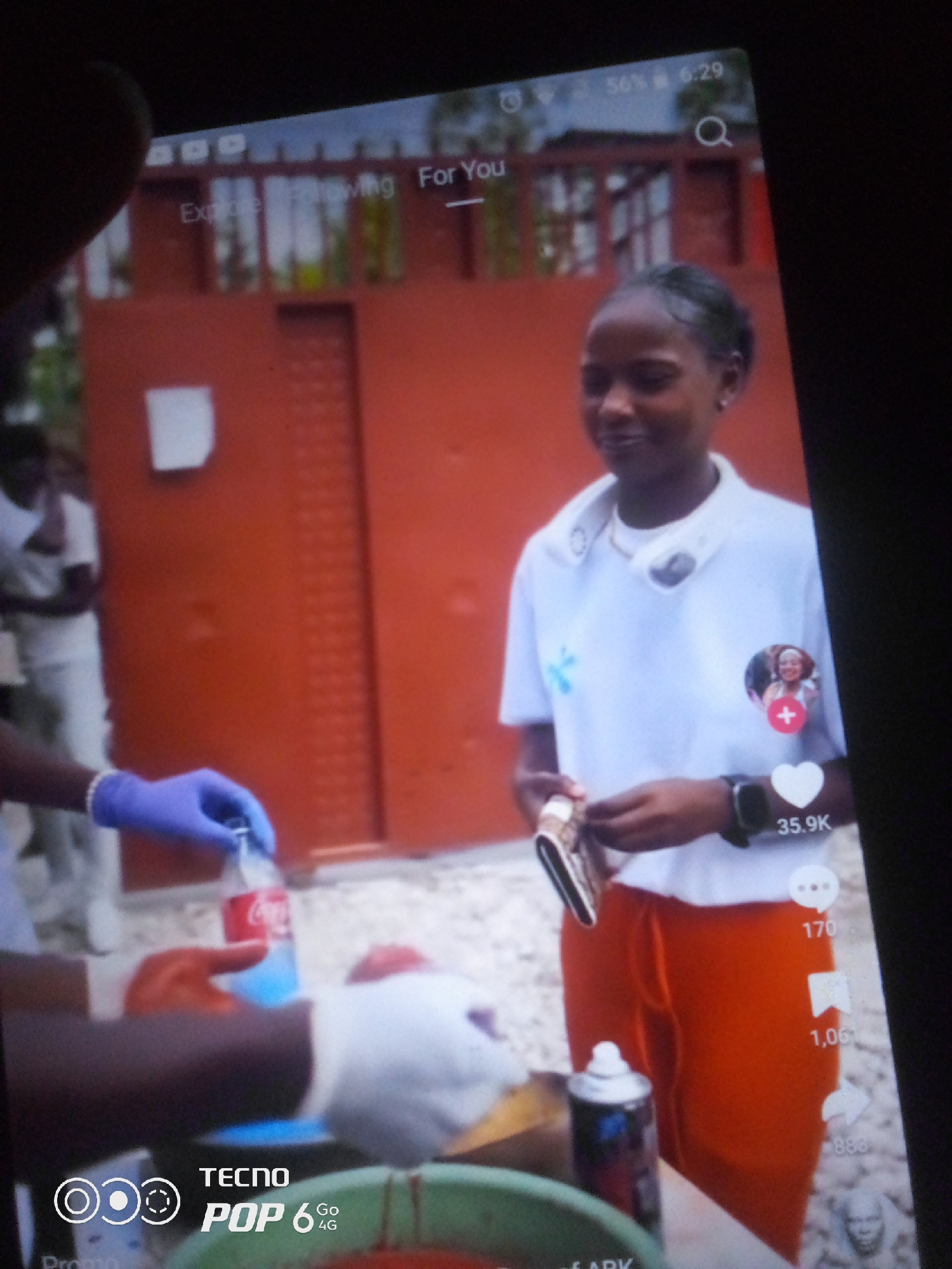 The image is a screenshot of a mobile phone screen displaying a social media video. The video shows a young Black female wearing a white t-shirt and orange pants, with a white neck fan around her neck and a watch on her left wrist. She is holding a wallet in her hands. To her right, social media interaction icons and counts are visible, including a heart with "35.9K" and other icons with numbers.
In the foreground of the video, a person wearing blue and white gloves is seen holding a red and white Coca-Cola bottle, appearing to pour liquid from it into a green container. Another person, partially visible, is wearing blue gloves. In the background, an orange-colored wall with dark vertical lines is present, and two other individuals in white shirts are visible.
The image also displays elements of the phone's user interface, including the time "6:29" and battery percentage "56%", as well as options like "Explore", "Following", and "For You". The bottom left corner of the screenshot shows "TECNO POP 6 Go 4G", indicating the phone model.
The location is not specified as Howayi, Nigeria.