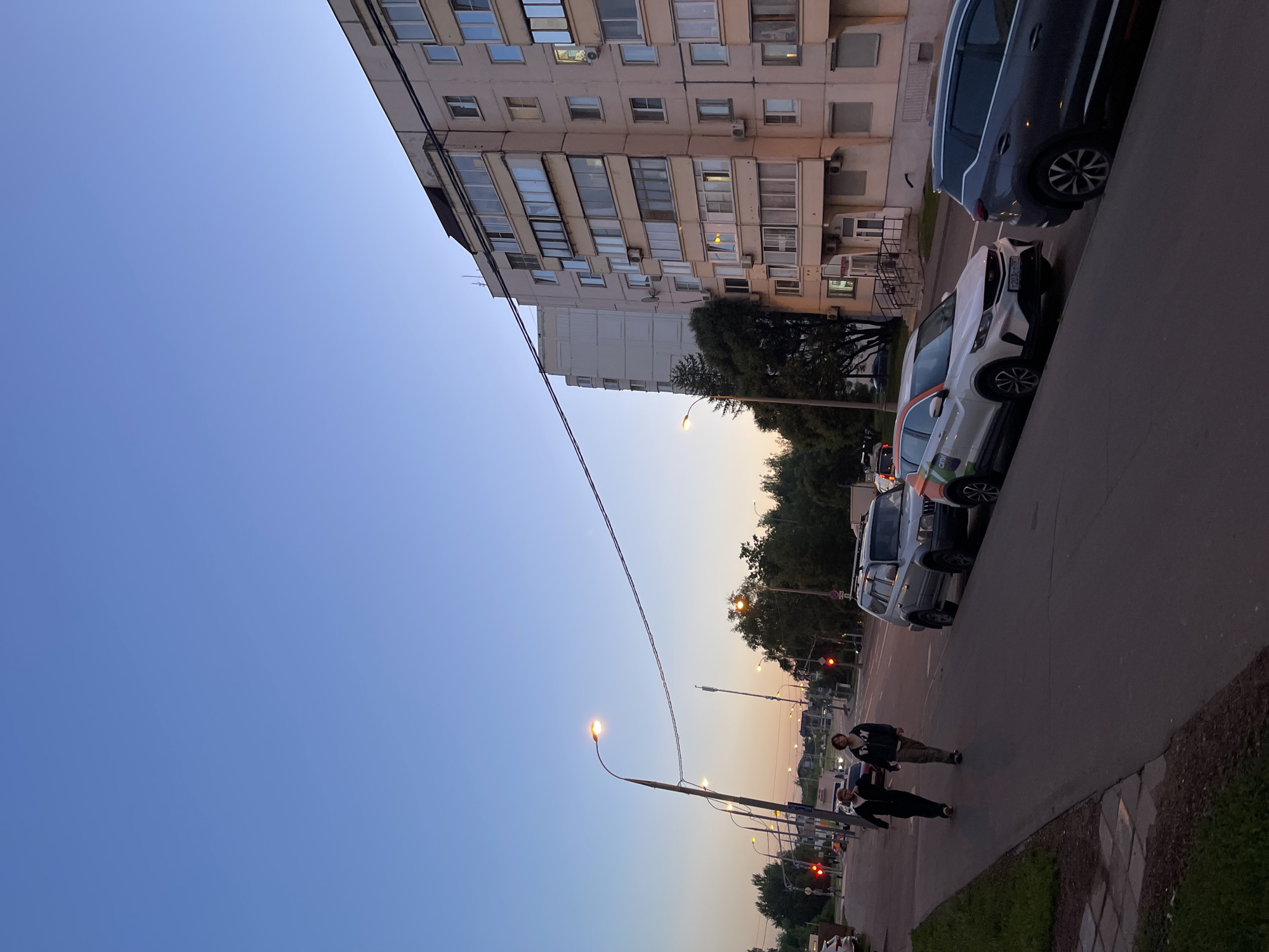 Two pedestrians walk on a sidewalk next to a street in Moscow, Russia, as dusk settles. A multi-story apartment building lines the right side of the street, with several cars parked along the curb. Traffic lights are visible in the distance, indicating ongoing activity despite the fading light. The sky is a clear, soft blue, transitioning to a pale orange near the horizon, suggesting the end of a day.